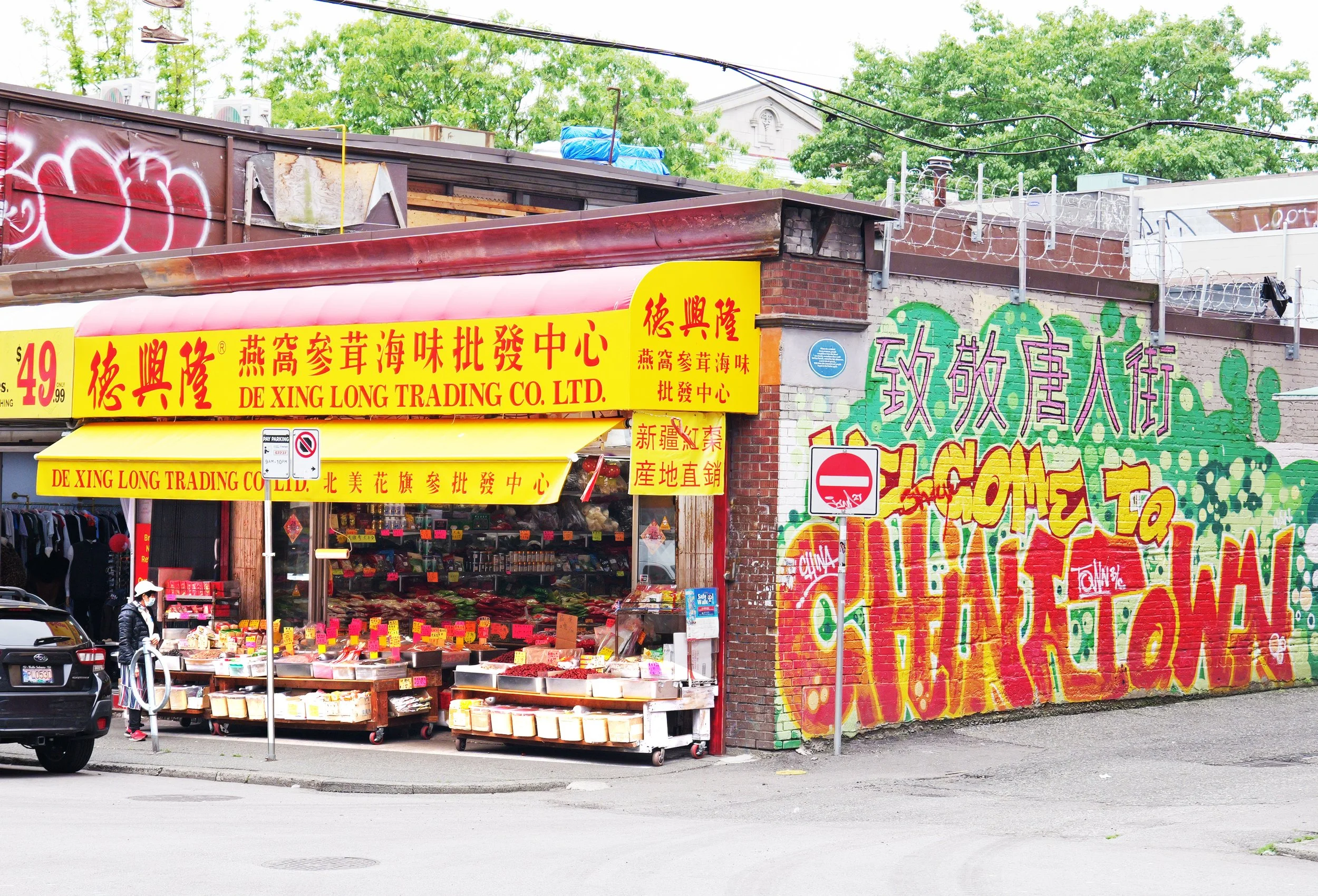 A street scene featuring a storefront with bright yellow signage in Chinese characters, selling seafood and produce. The store has an open front with visible fruits, vegetables, and seafood displayed, and there are pedestrians, cars, and a colorful graffiti mural on the brick wall next to the store.