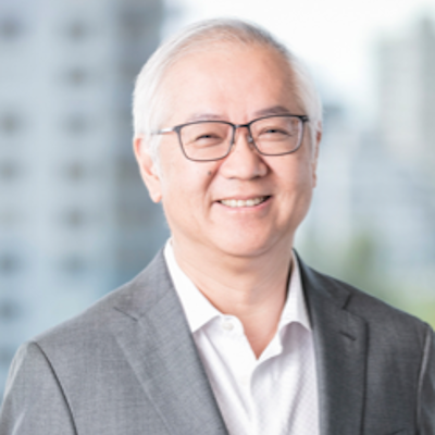 Smiling older man in glasses wearing a gray suit and white shirt, standing in front of a blurred cityscape background.