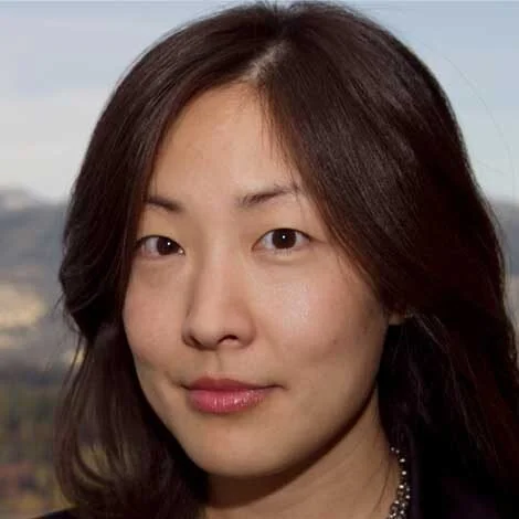 A woman with straight, shoulder-length dark hair and light makeup, outdoors with a blurred cityscape background.