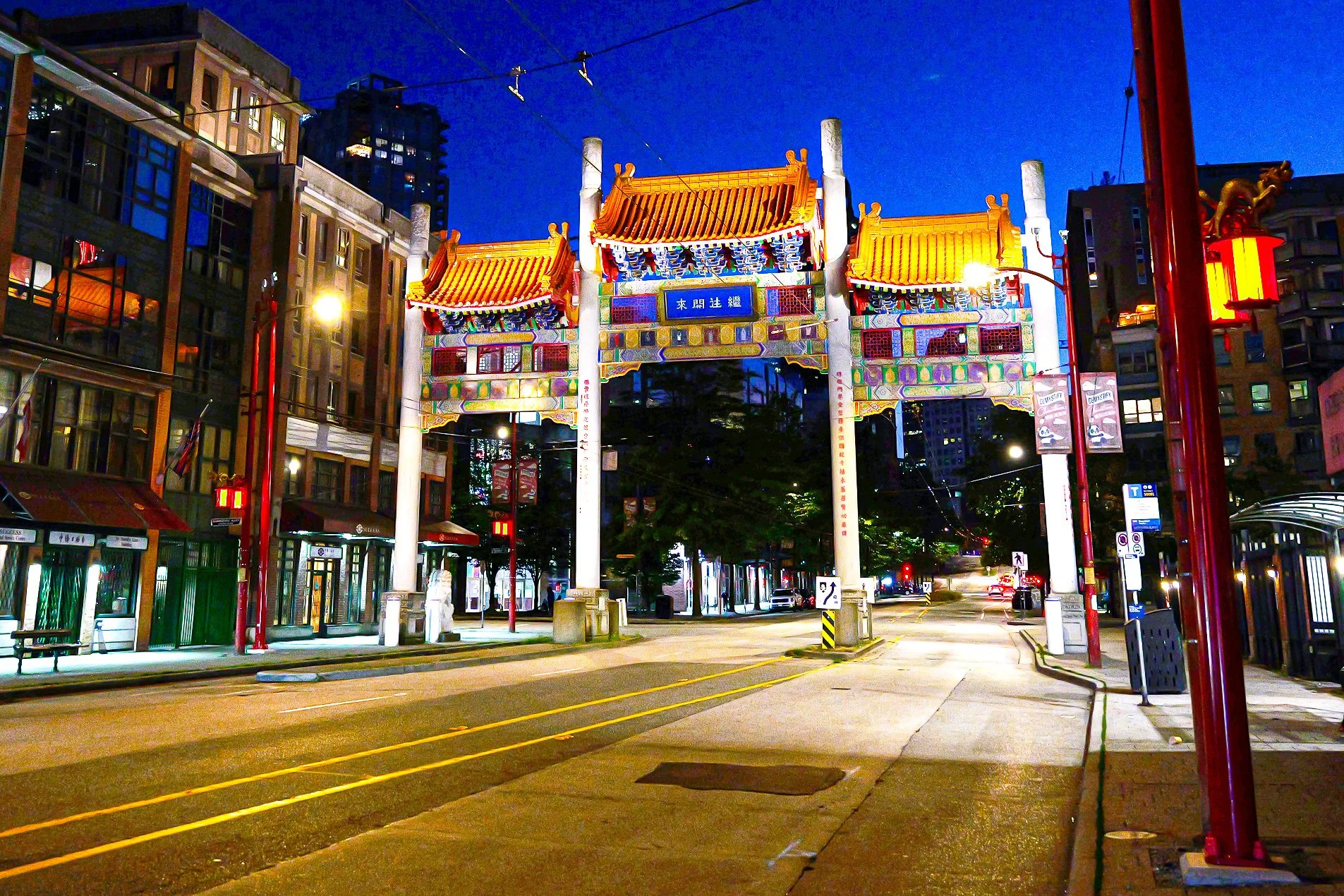 Night view of a traditional Chinese style gate with yellow-tiled roofs, colorful decorative elements, and Chinese characters on a blue sign. City buildings and street with few cars and streetlights in the background.