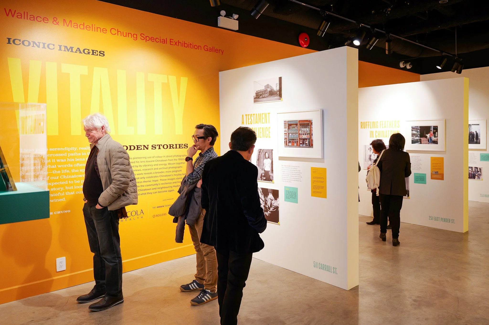 People viewing photographs and information panels at an art exhibition in a gallery with orange and white walls.