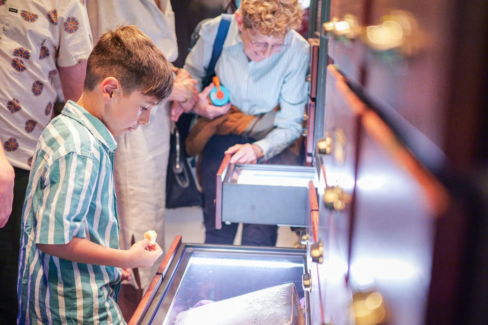 A young boy with a striped shirt looking at an exhibit case with artifacts, while an older woman with curly hair and glasses looks at the same display.