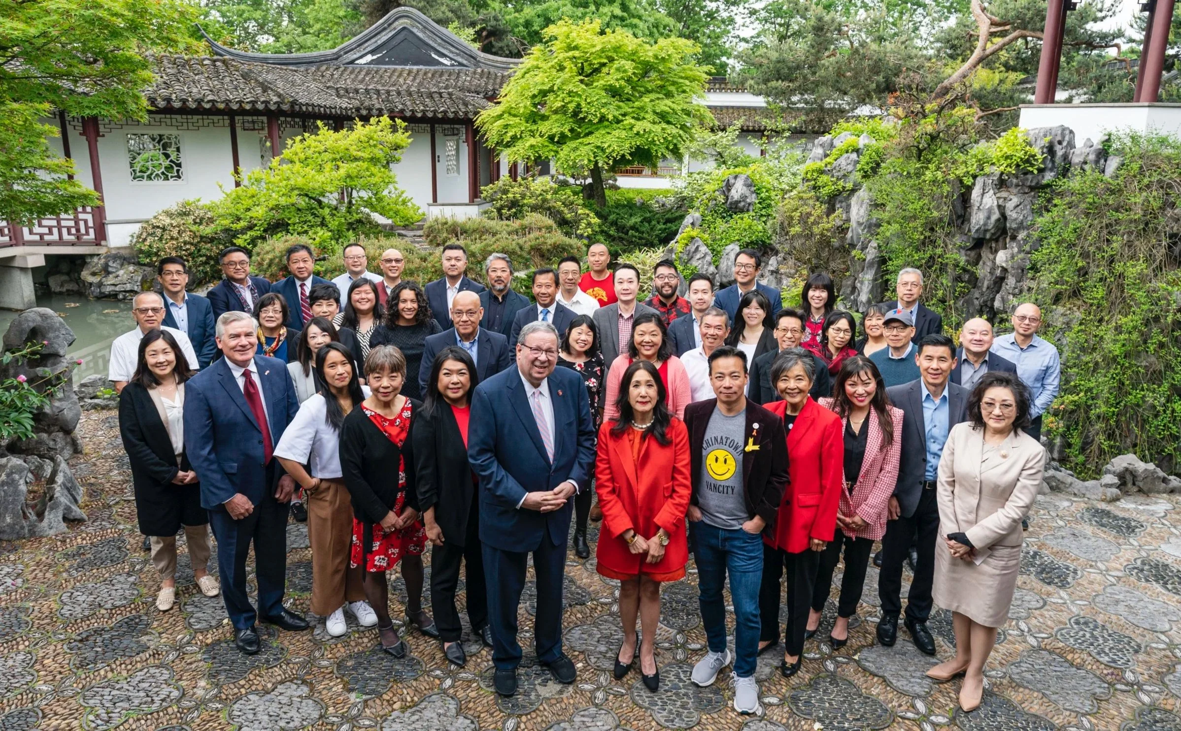 Group of diverse people standing outdoors in front of a traditional Asian-style building with greenery and rocks.