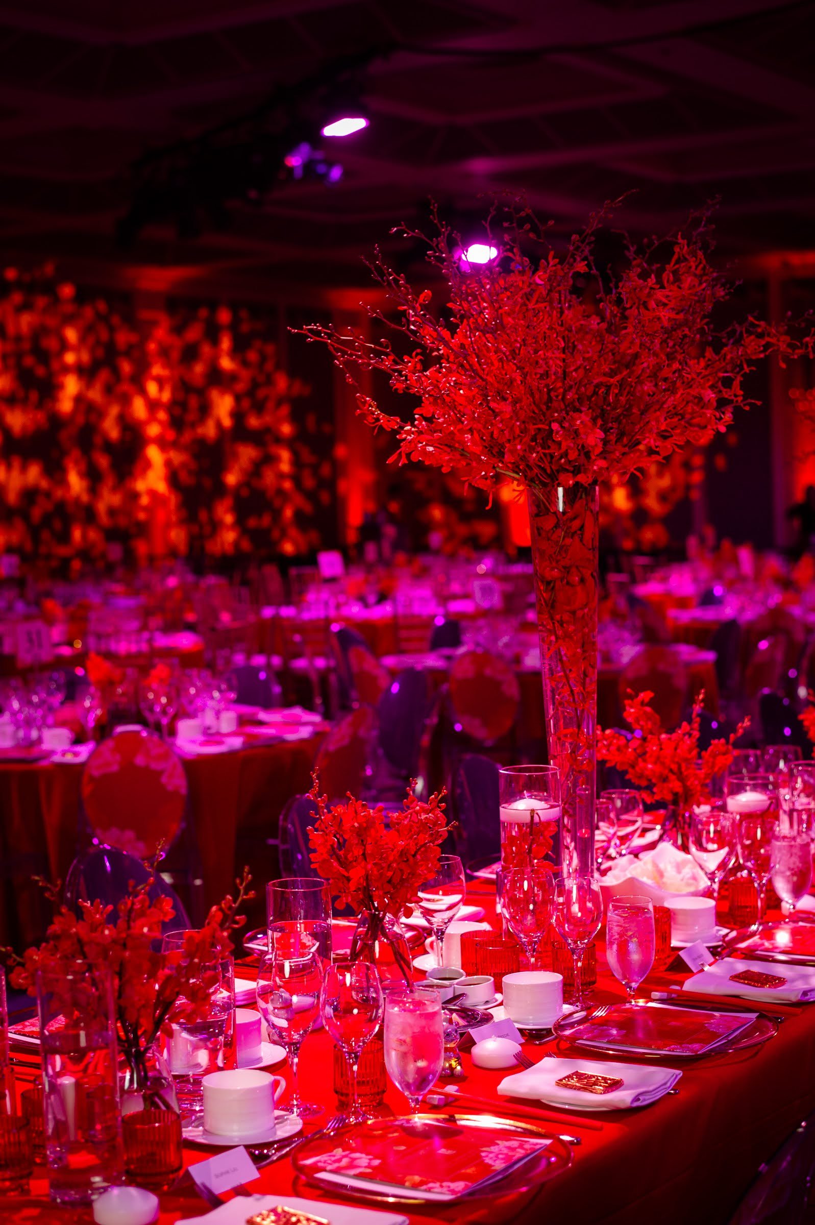 Elegant banquet hall decorated with red lighting, featuring a tall floral centerpiece with red flowers on a table set with glassware, plates, and candles.