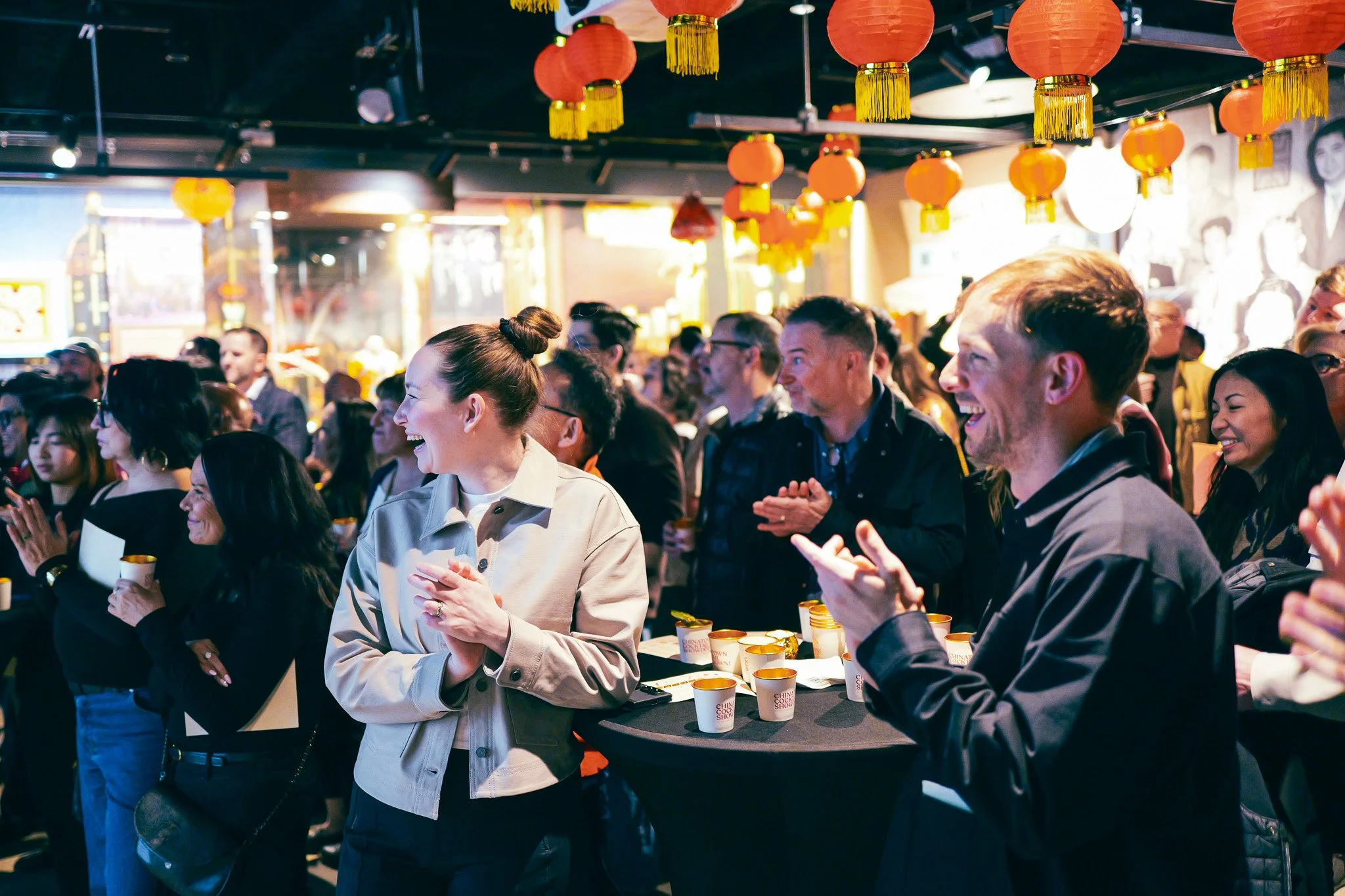 A group of people gathered at an indoor event with Chinese New Year decorations, including red lanterns hanging from the ceiling. The attendees are smiling, clapping, and holding cups, indicating a celebratory atmosphere.