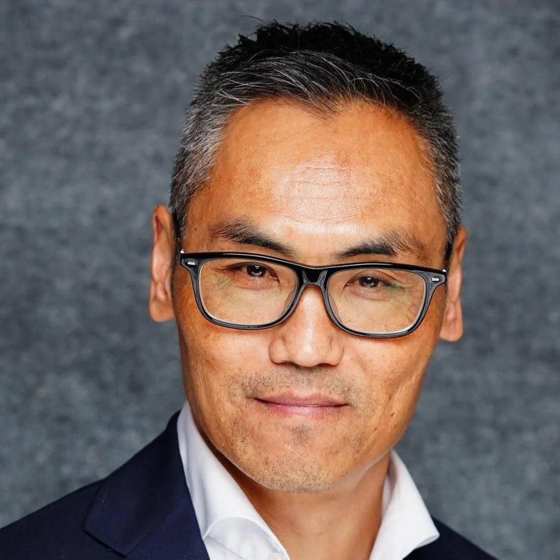Close-up portrait of a middle-aged man with short, salt-and-pepper hair, wearing black-rimmed glasses and a dark suit with a white dress shirt, against a gray textured background.