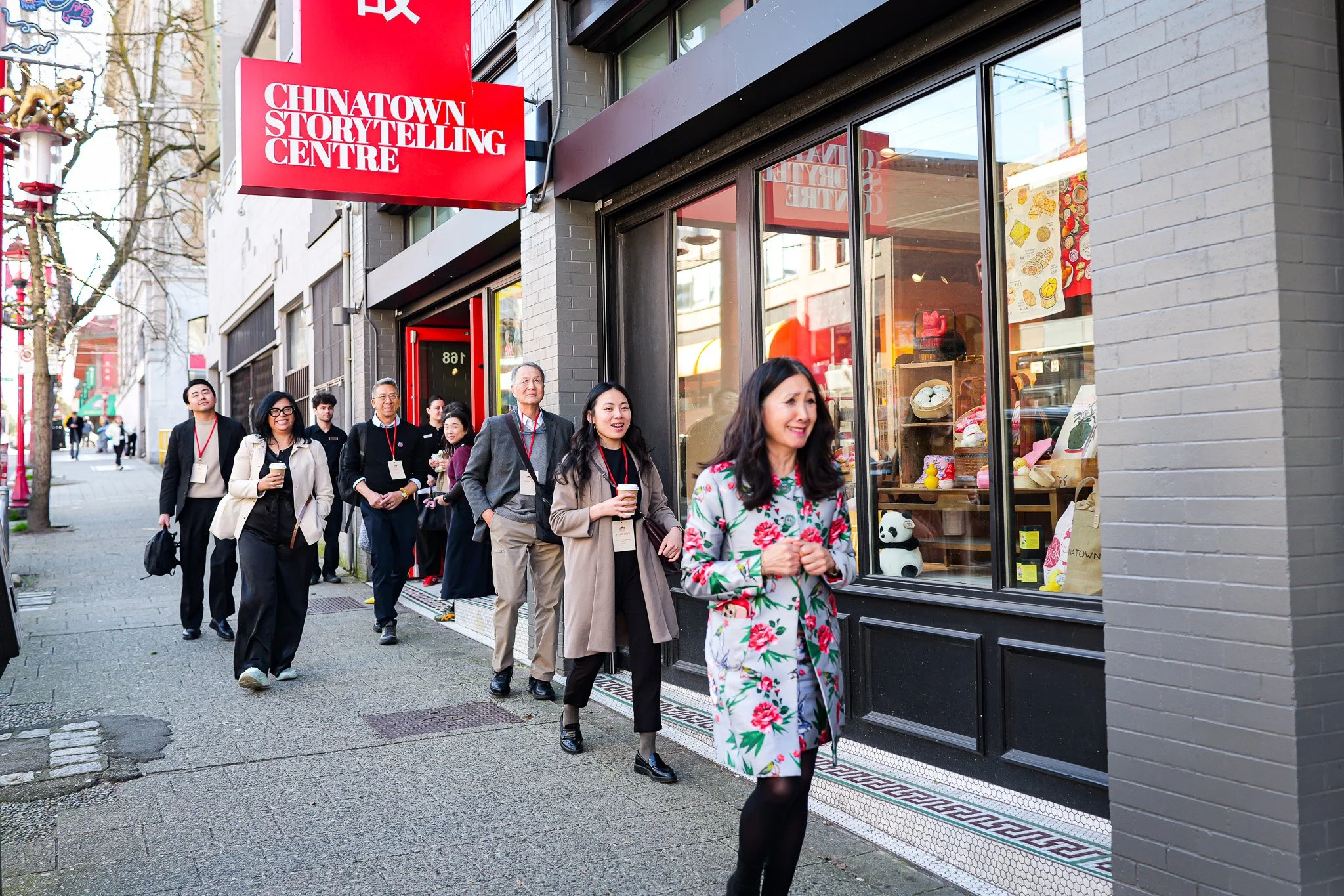 People standing in line outside the Chinatown Storytelling Centre in an urban area with brick buildings and street trees.
