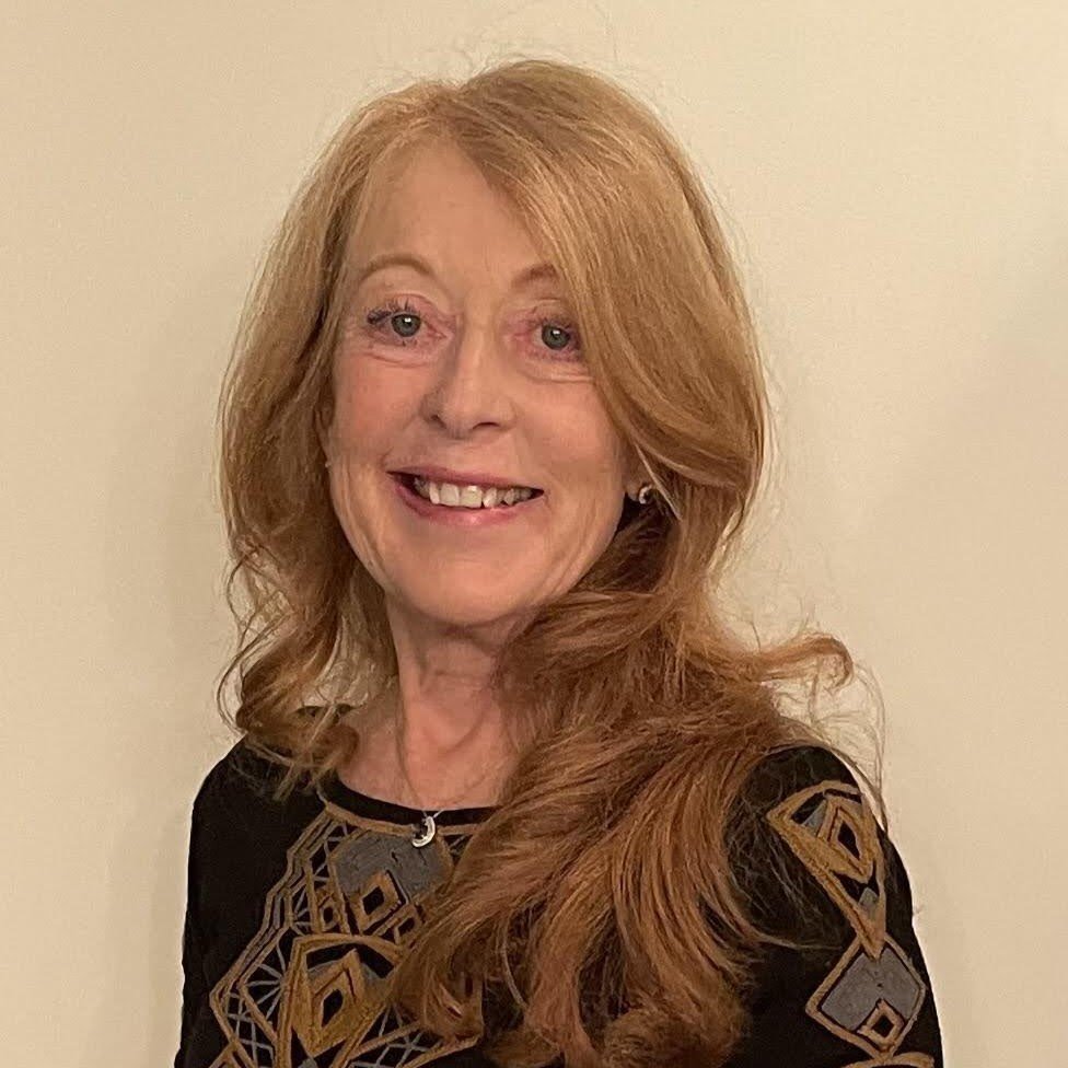 A woman with long, wavy red hair and light skin, smiling, wearing a black top with a geometric pattern and a necklace, standing against a white background.
