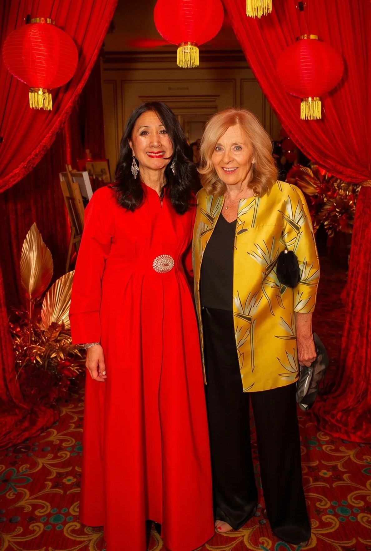 Two women standing together at a festive event with red lanterns and curtains in the background. One woman is wearing a red dress with jewelry, and the other is in a yellow jacket with a black top and pants, smiling at the camera.