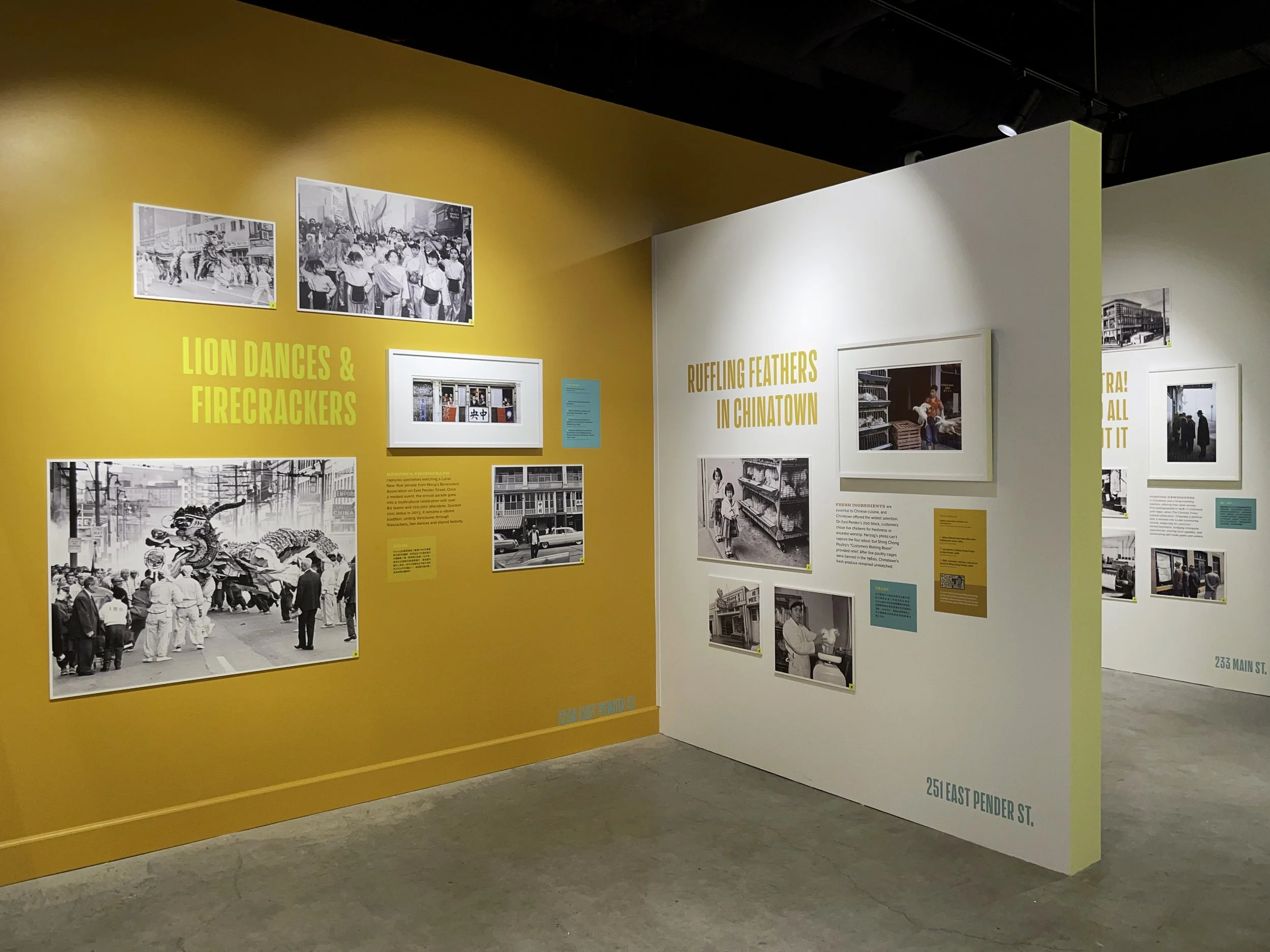 Exhibit display with black and white historical photographs and colorful text panels about Chinatown, including photos of lion dance performances, firecrackers, and people shopping or working, with orange and white walls and small green and blue informational labels.