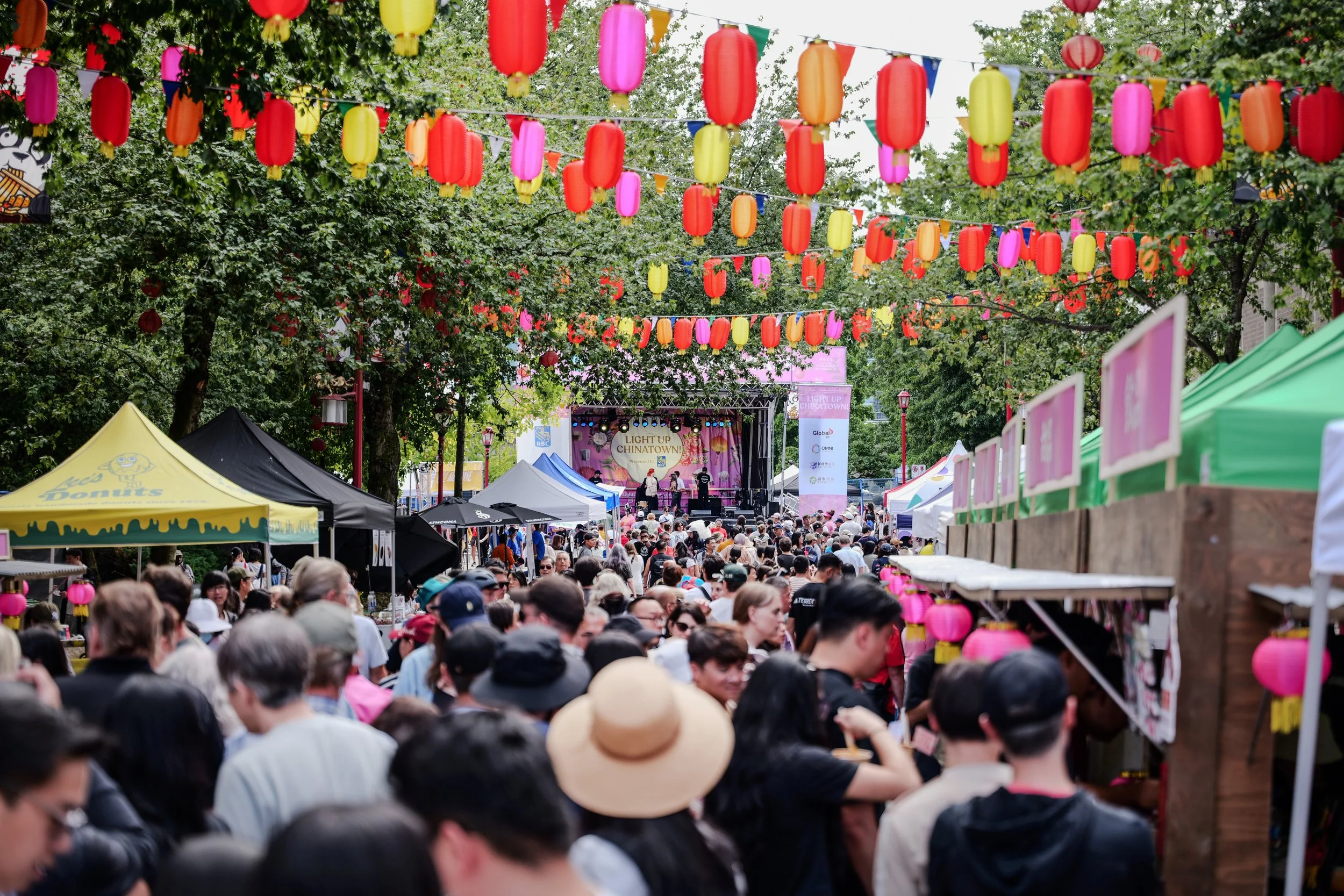 Crowd attending an outdoor festival with colorful lanterns and banners, stage with performers in the background, trees, and vendor tents.
