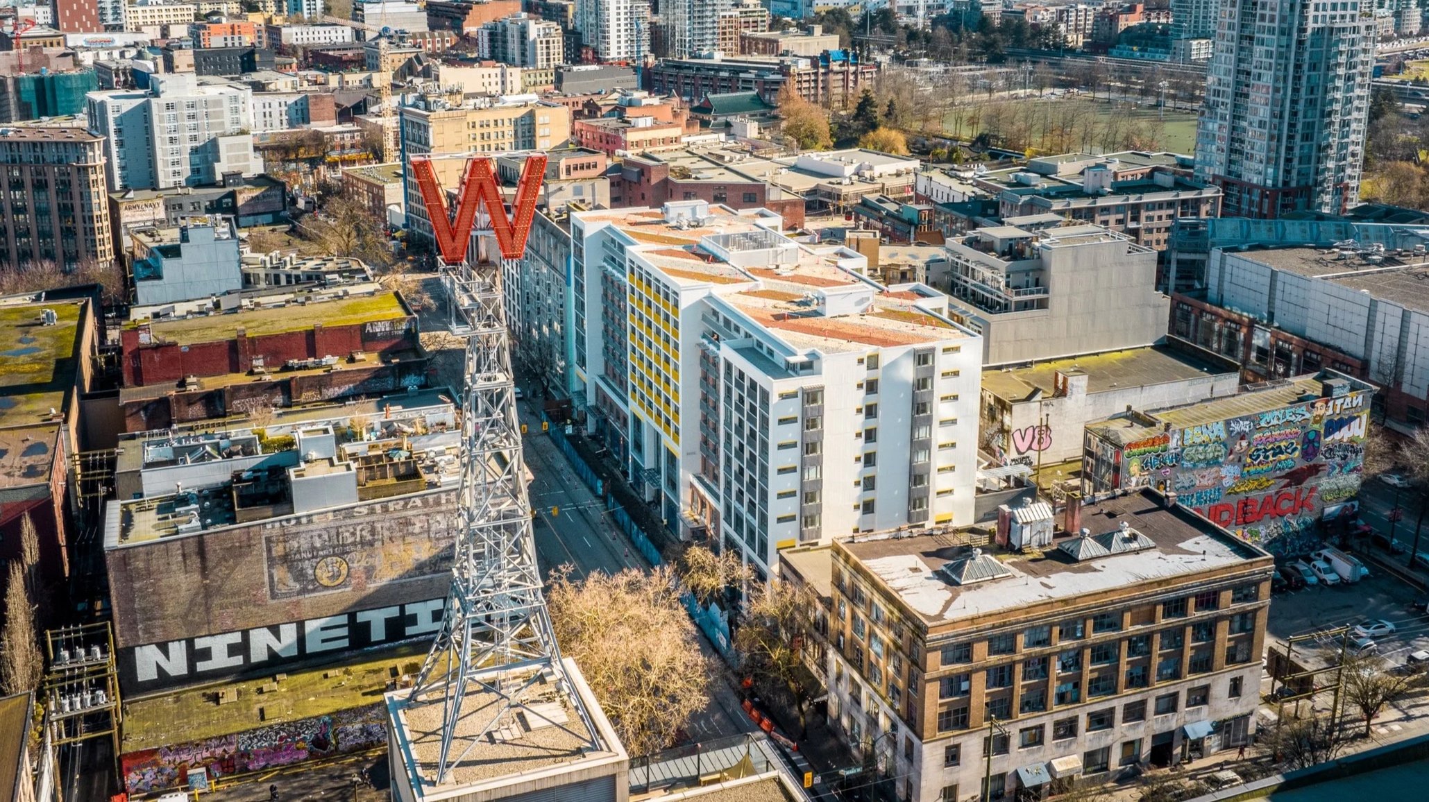 An aerial view of a cityscape showing numerous buildings, high-rise apartments, and a construction crane with a red 'W' logo, amidst street views, graffiti-covered walls, and a park with trees in the background.