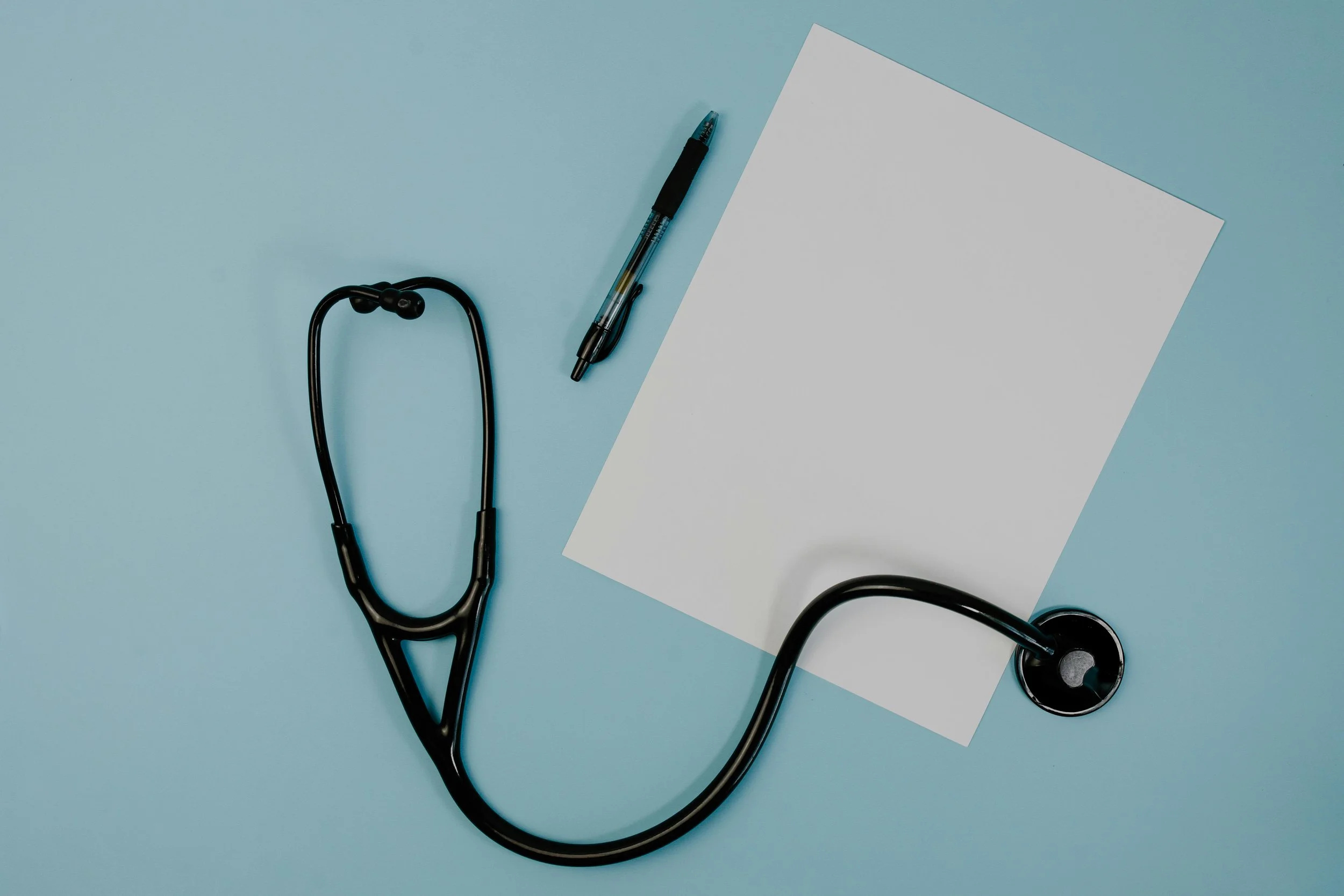 A stethoscope, a black ballpoint pen, and a blank white sheet of paper on a light blue background.