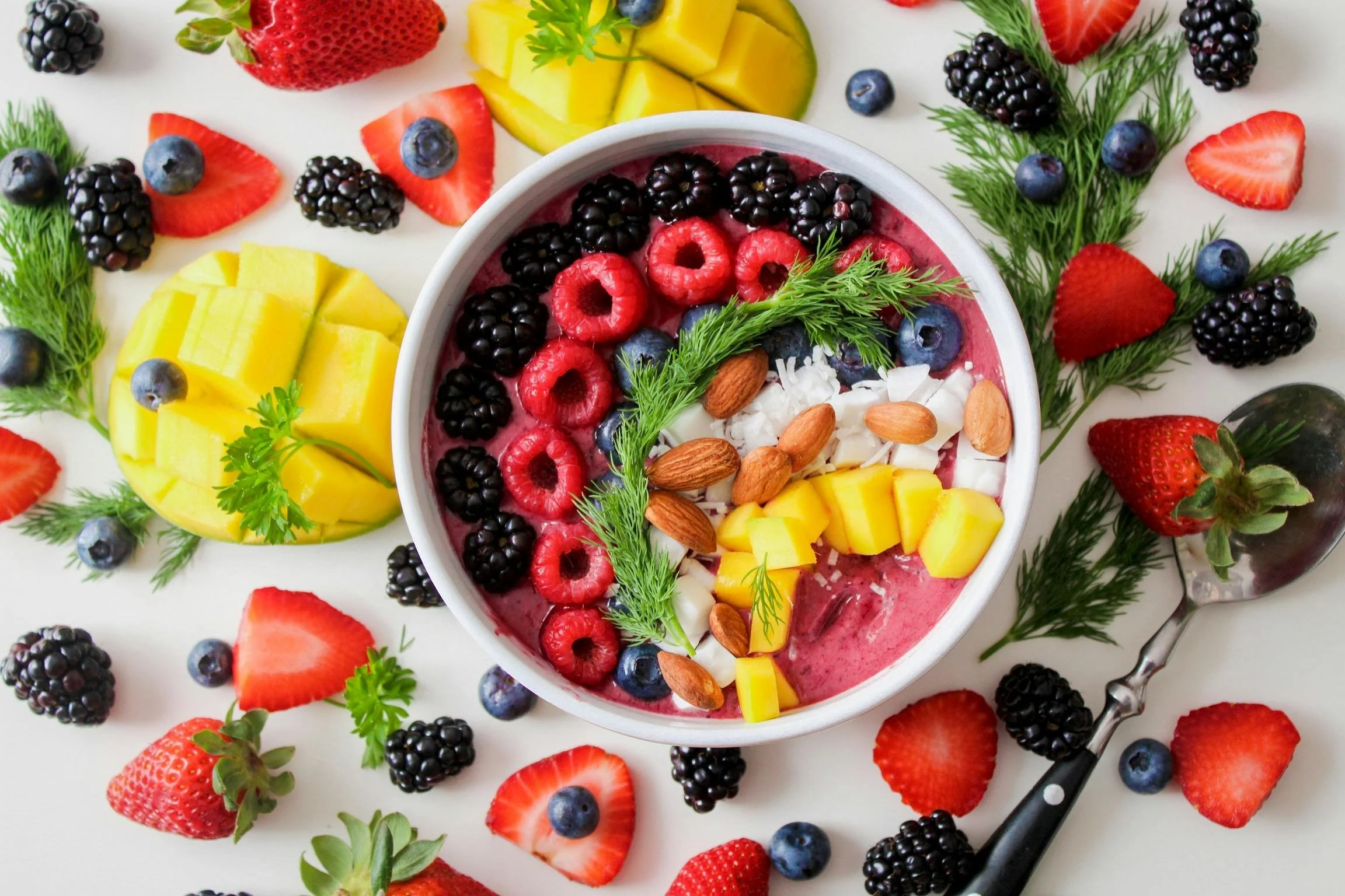 A bowl of mixed berry smoothie topped with raspberries, blackberries, blueberries, chopped almonds, shredded coconut, and small mango cubes, surrounded by scattered strawberries, blackberries, blueberries, and sprigs of green herbs on a white surface.