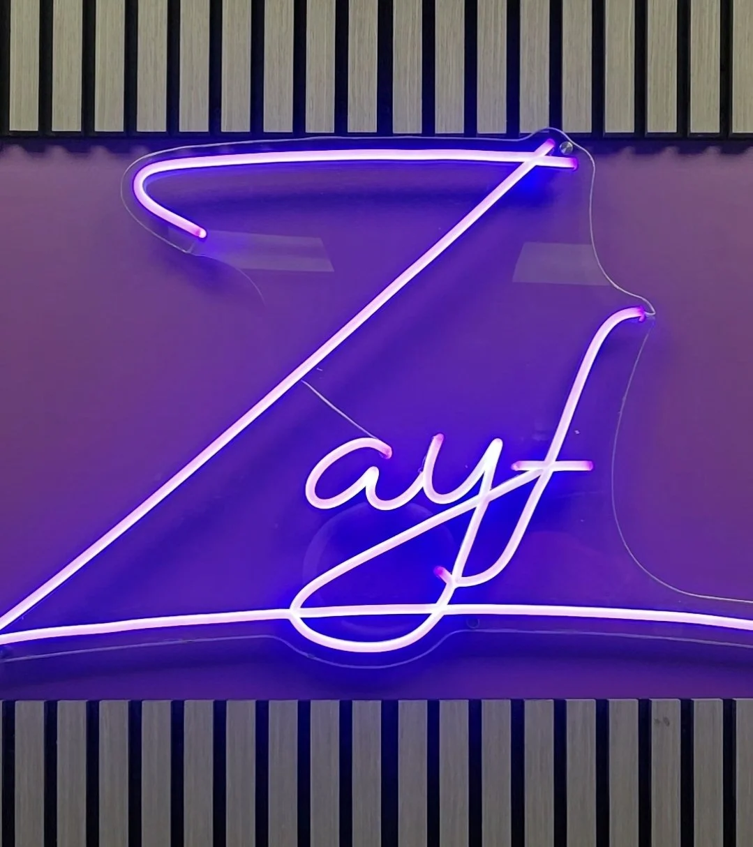 Neon sign reading 'Zayf' with purple and pink colors, mounted on a dark wall with wooden paneling at the top and bottom.