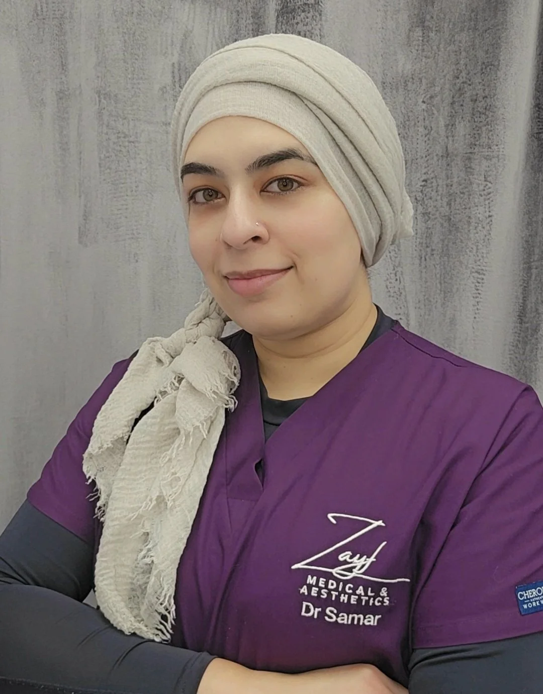 A woman, Dr Samar, wearing a beige headscarf and purple medical scrubs with the logo 'Zayf Medical & Aesthetics' and the name 'Dr. Samar', standing against a gray background.