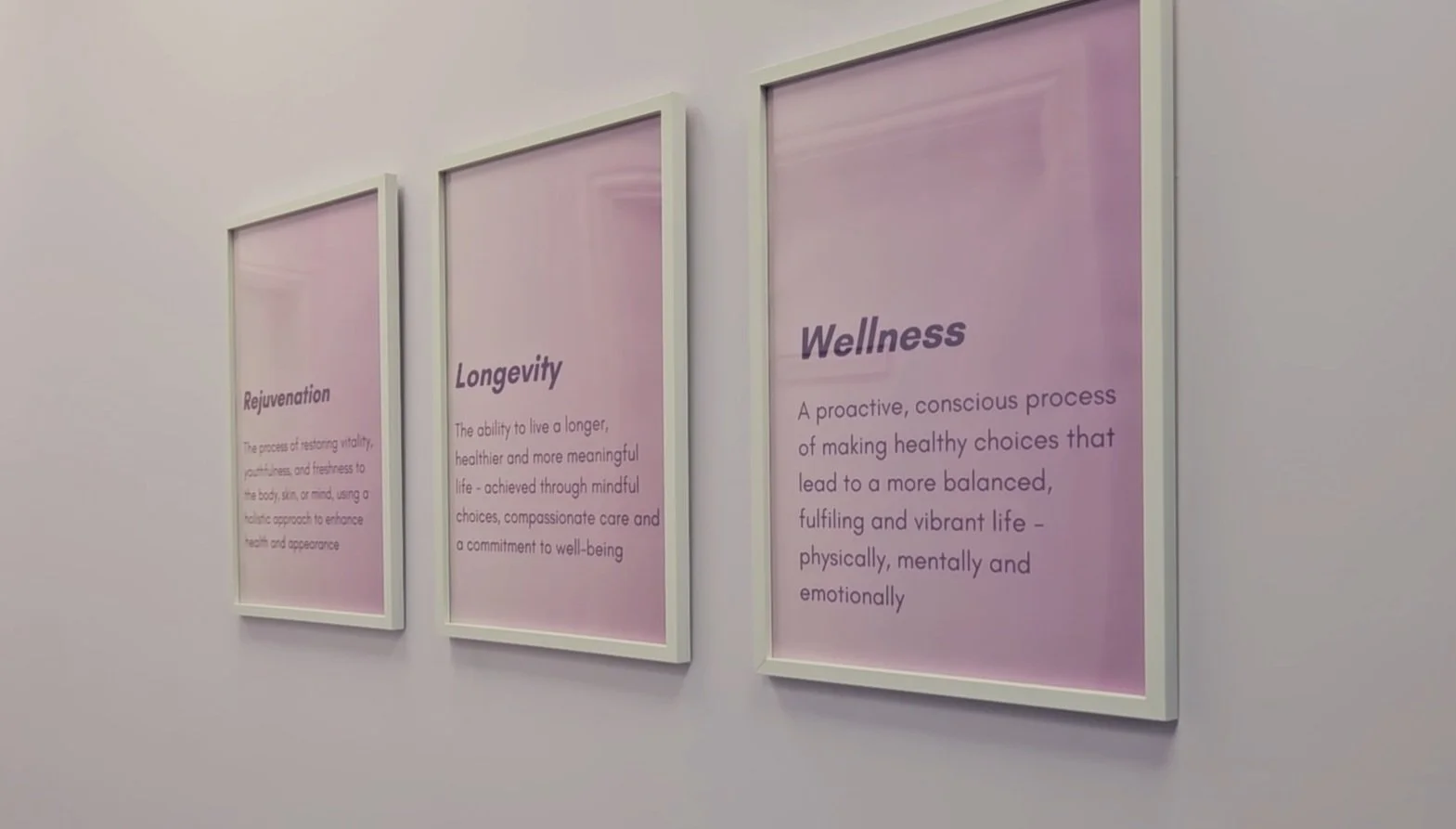 Three framed signs on a white wall with pink backgrounds and purple text, titled Rejuvenation, Longevity, and Wellness, describing health and wellbeing concepts.