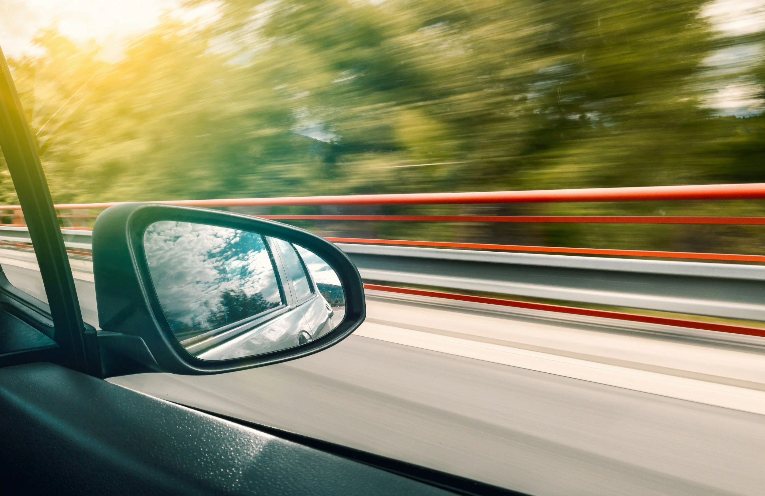 A moving view from inside a vehicle showing the side mirror and highway railing, with blurred trees and sky in the background.