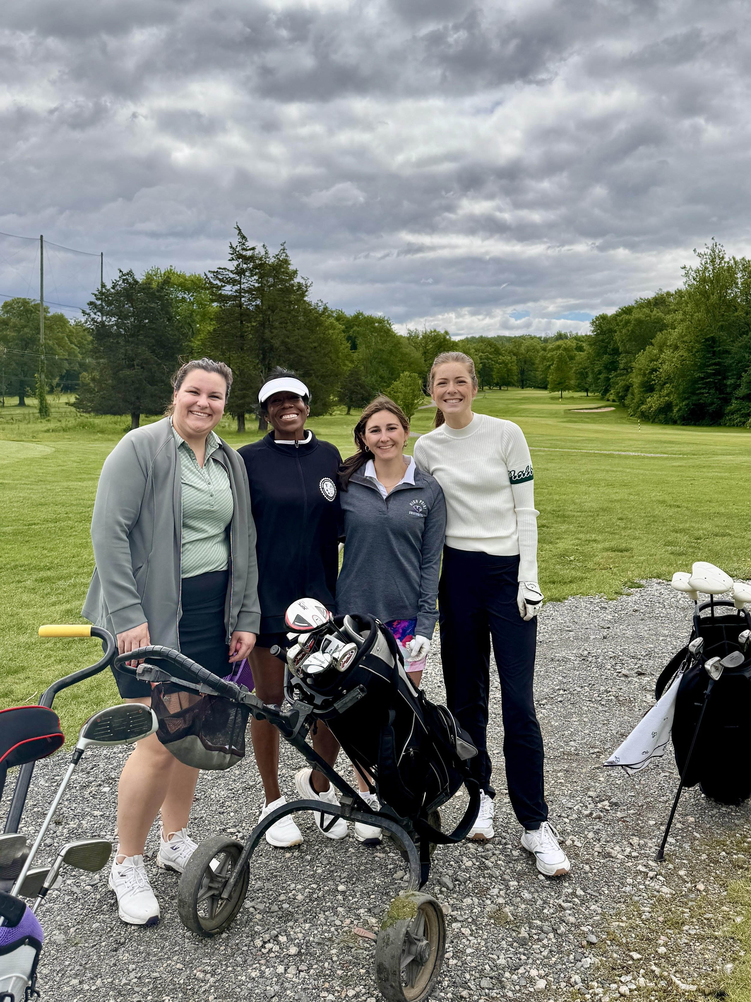 Four women standing on a golf course with golf clubs and carts, smiling, during a cloudy day.