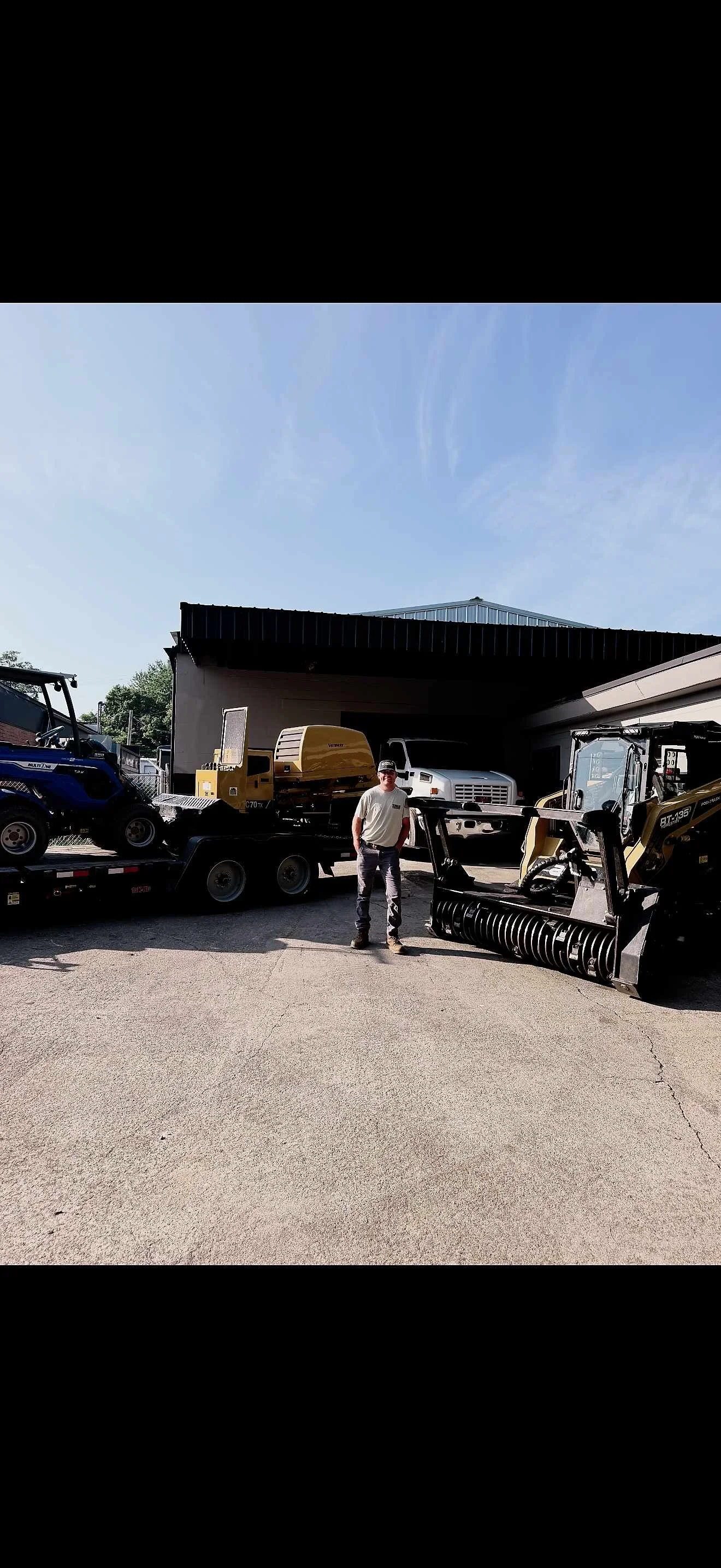 A man standing in front of various construction and landscaping equipment, including a skid steer loader, a small trailer with equipment, and a white truck, outside a garage with a black roof under a clear blue sky.