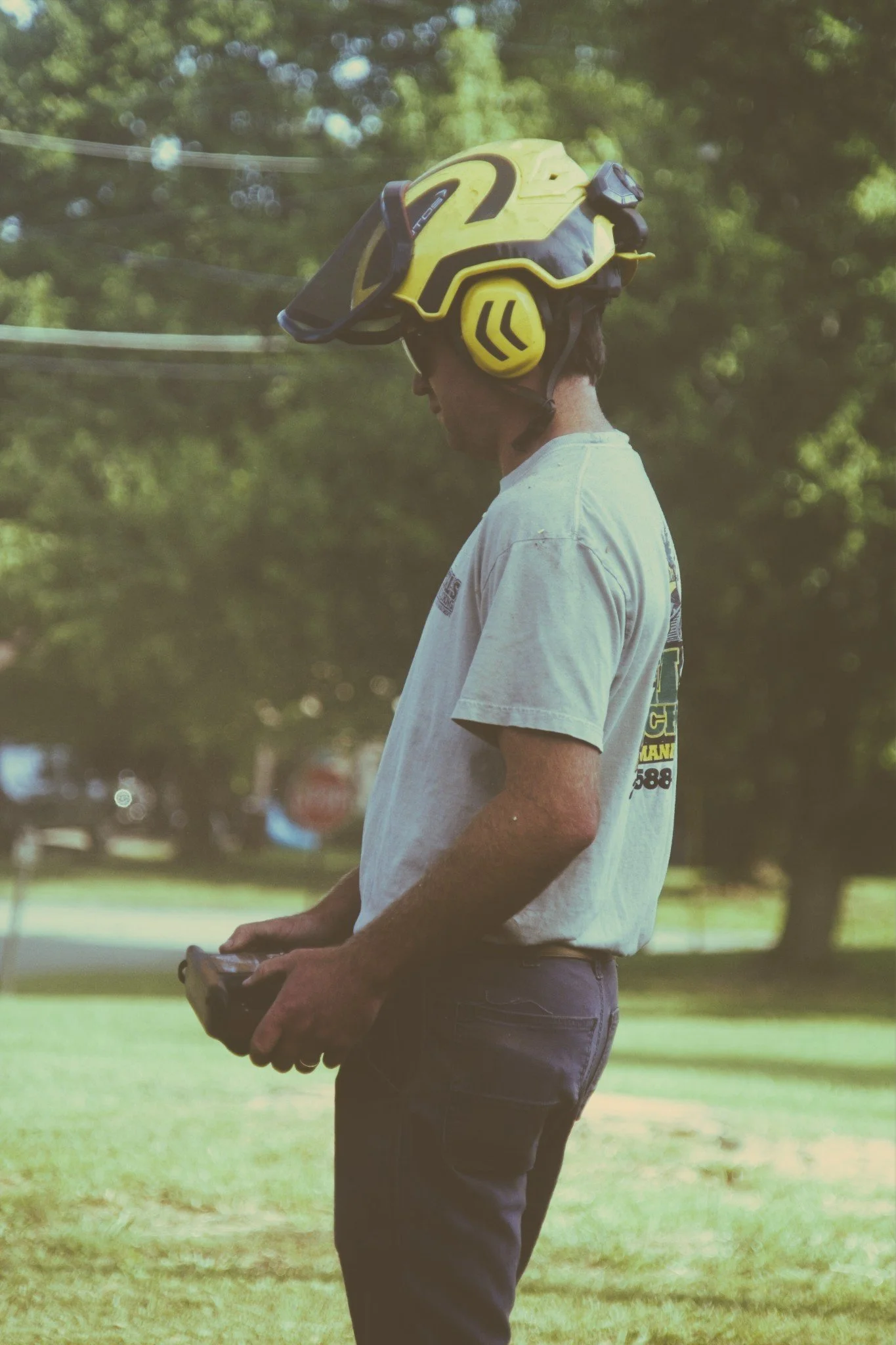 A man standing outdoors in a park with trees in the background, wearing a helmet and casual clothes, holding a remote control.