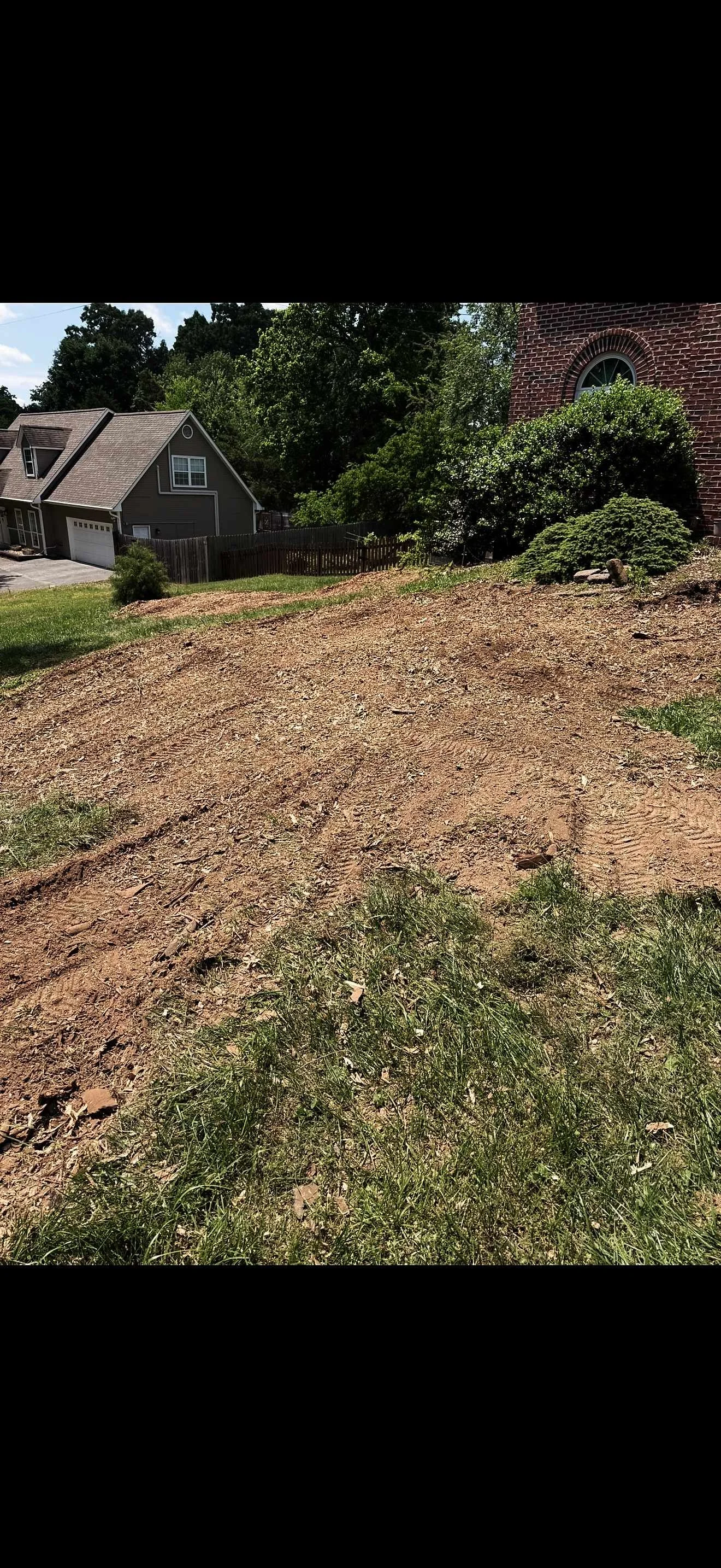 A dirt slope with tire tracks, bordered by grass and bushes, in a residential area with houses and trees in the background.