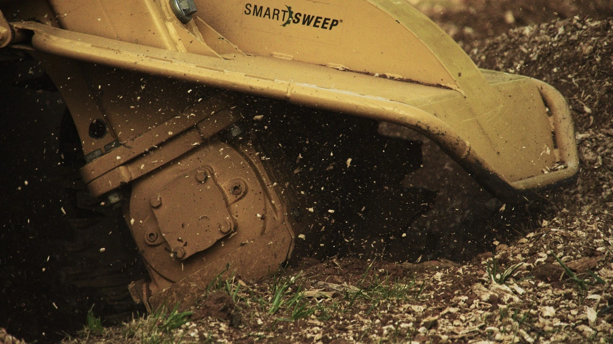 Close-up of a yellow robotic stump grinder on the ground, covered in dirt and grass clippings, with soil and small plants nearby.