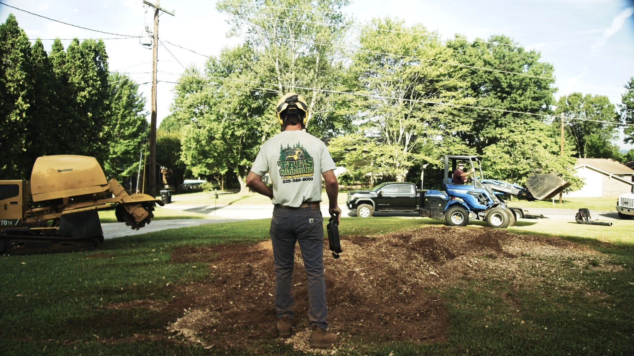 A man wearing a helmet and Talis Mulching shirt stands on a lawn in a neighborhood, overseeing landscaping work, with construction equipment, a blue utility vehicle, and a black pickup truck nearby.