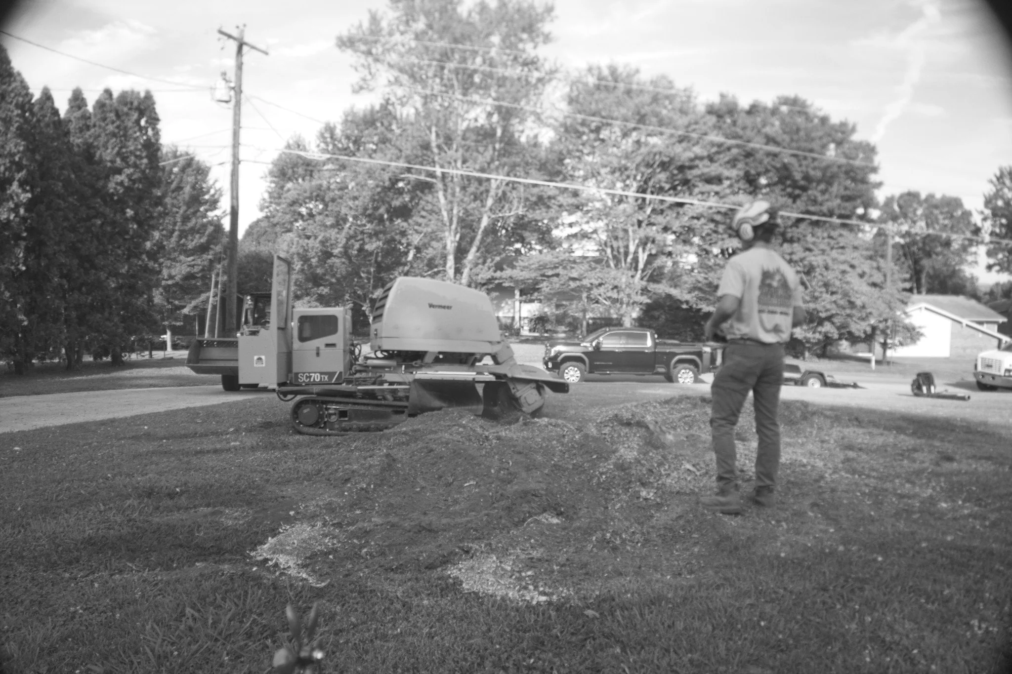 A worker standing outdoors on a grassy area near a machine, with trees and a few vehicles in the background, in a black-and-white photo.