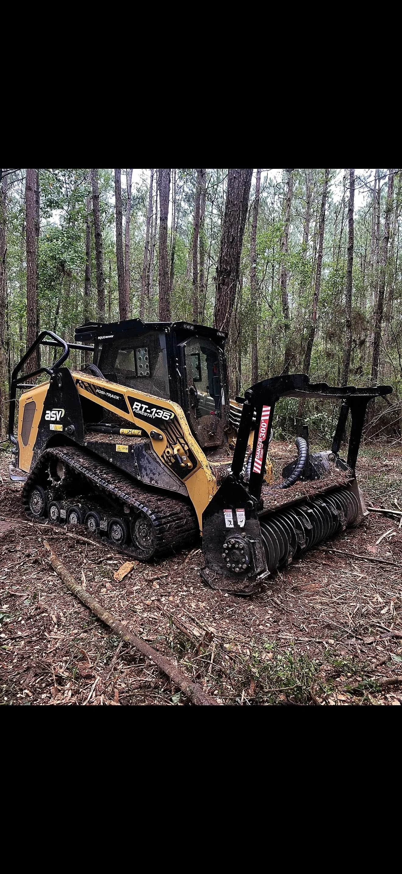 A Skid steer loader with a mulcher attachment is in a forested area, clearing fallen branches and debris from the ground.