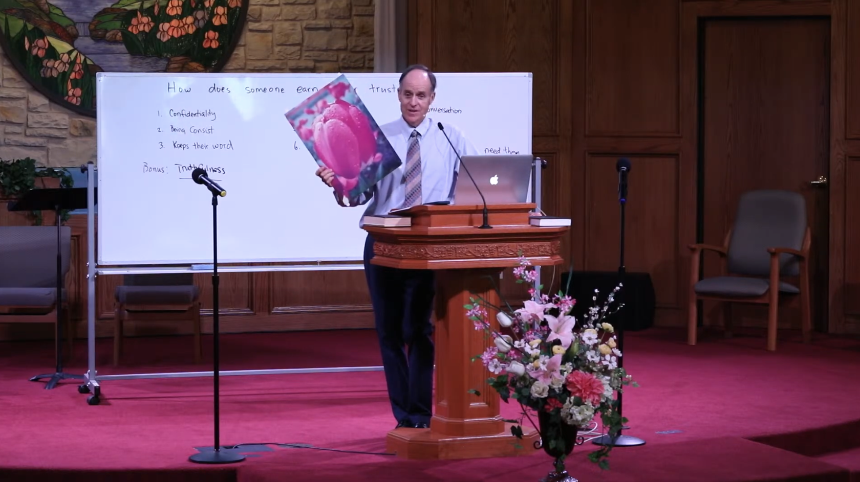 A man in a dress shirt, tie, and dark pants stands at a wooden pulpit in a church or conference room, holding a framed picture of pink flowers. Behind him, a whiteboard with handwritten notes about earning trust, a laptop, and a microphone on a stand. There are chairs, a floral arrangement, and decorative wall art including a stained glass window in the background.