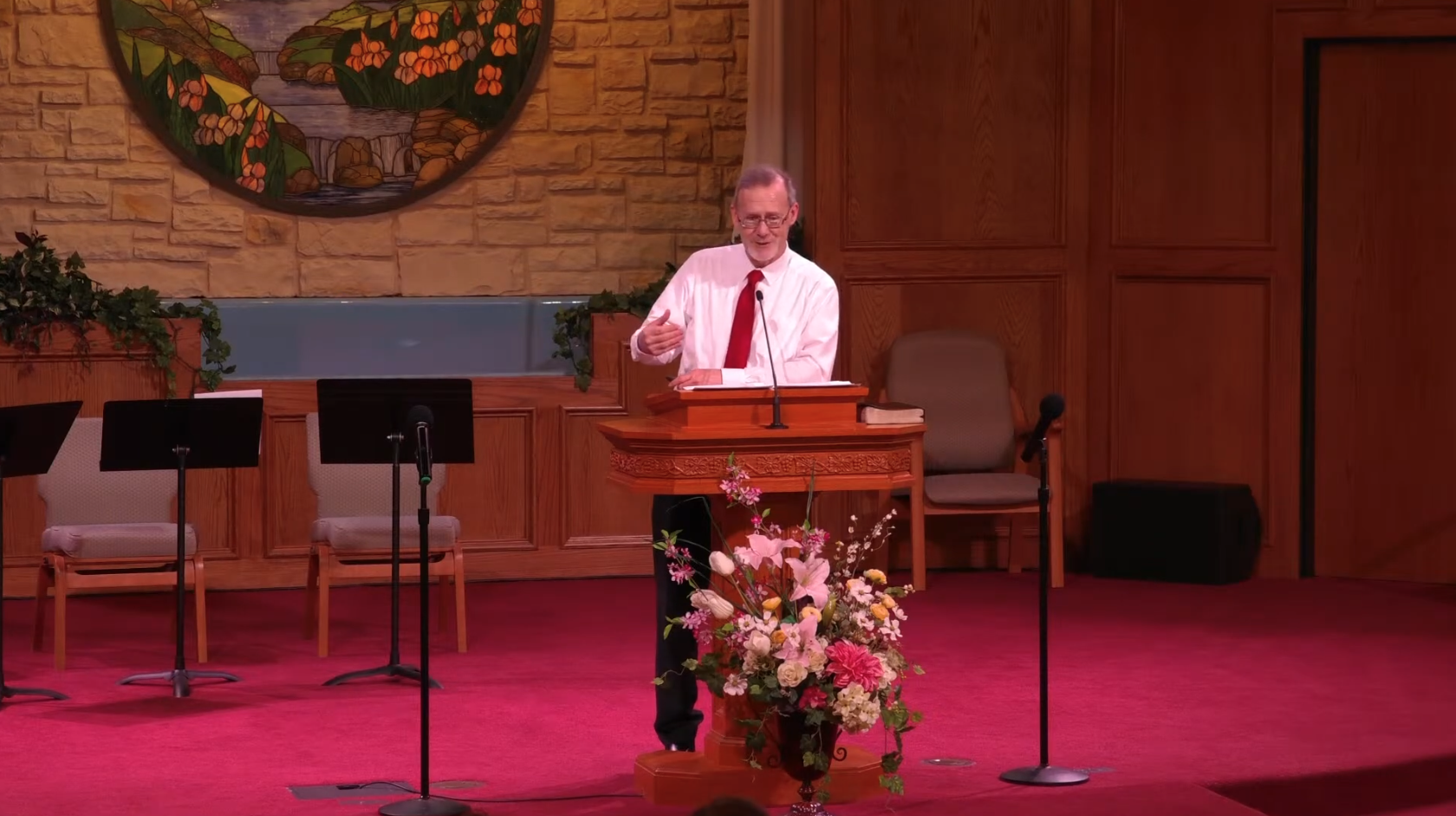 A man with glasses and gray hair, wearing a white shirt and red tie, speaking at a wooden podium decorated with a large flower arrangement, in a church or auditorium with wood-paneled walls and a stone backdrop.
