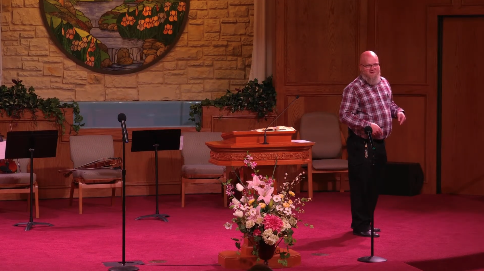 A man with a bald head and beard wearing glasses, a red plaid shirt, and black pants standing on a stage, speaking or gesturing, with a microphone in front of him. The stage has wooden walls and a brick wall background with a large circular stained-glass artwork featuring flowers and nature scene. There are chairs, music stands, a floral arrangement in the foreground, and decorative green plants.