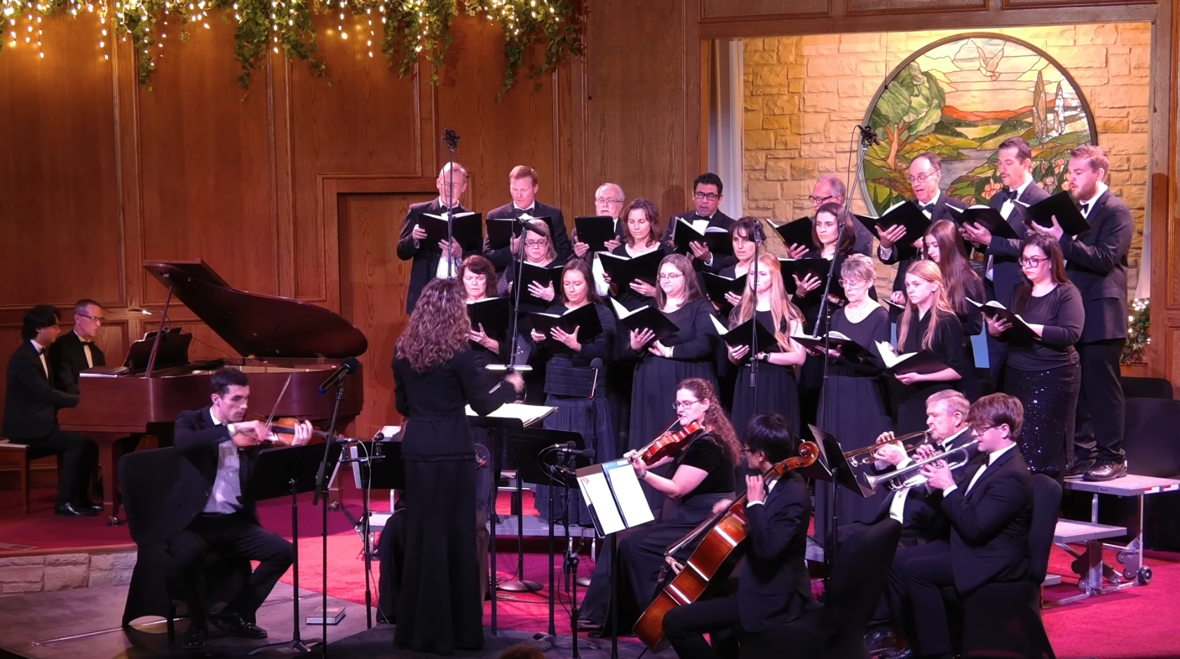 A choir of men and women dressed in formal black attire singing on a stage with a conductor, accompanied by a small orchestra including a pianist, violinist, cellist, and trumpeter, in a room with wood-paneled walls and holiday decorations.
