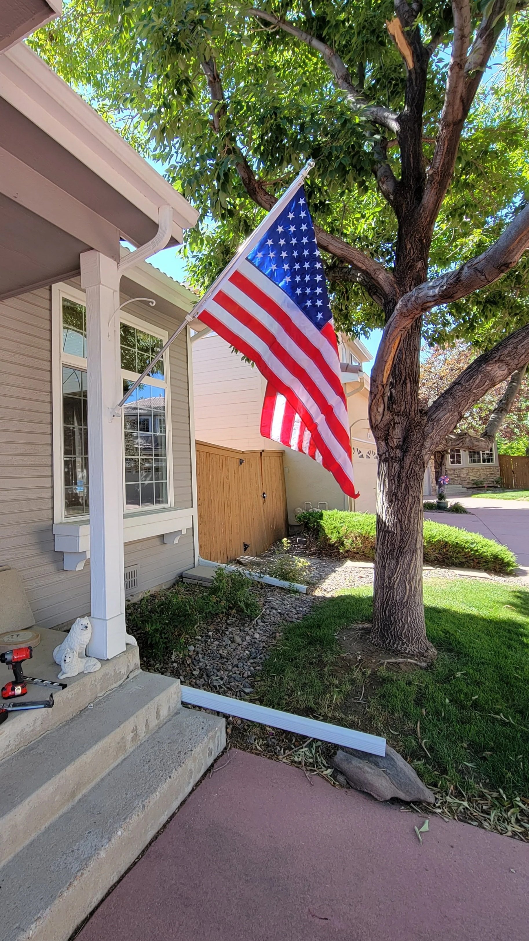 American flag hanging on a pole outside a house next to a tree, with green leaves and a bright sky in the background.