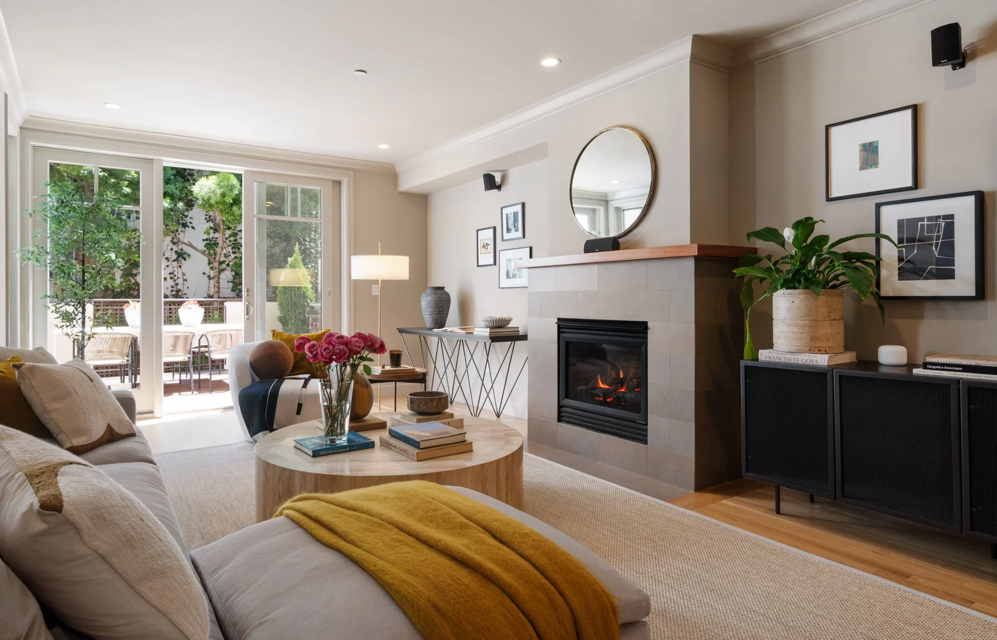 Living room with a cozy fireplace, large windows with a view of greenery, a round coffee table with books and a vase of pink roses, framed artwork on the wall, and a mix of modern and traditional decor.