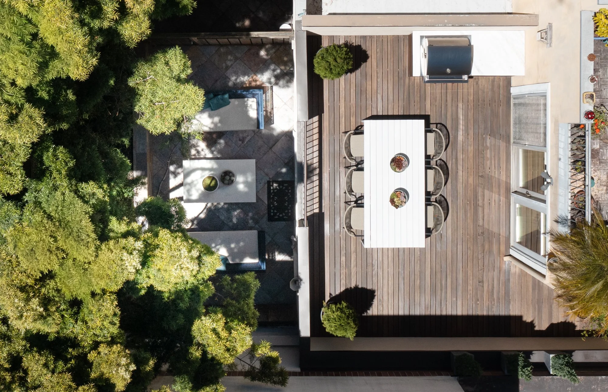 Overhead view of a wooden outdoor patio with a white dining table, six chairs, and two potted plants. To the left, dense green foliage and trees shade part of the patio. To the right, a windowed wall with some outdoor decorations and plants.