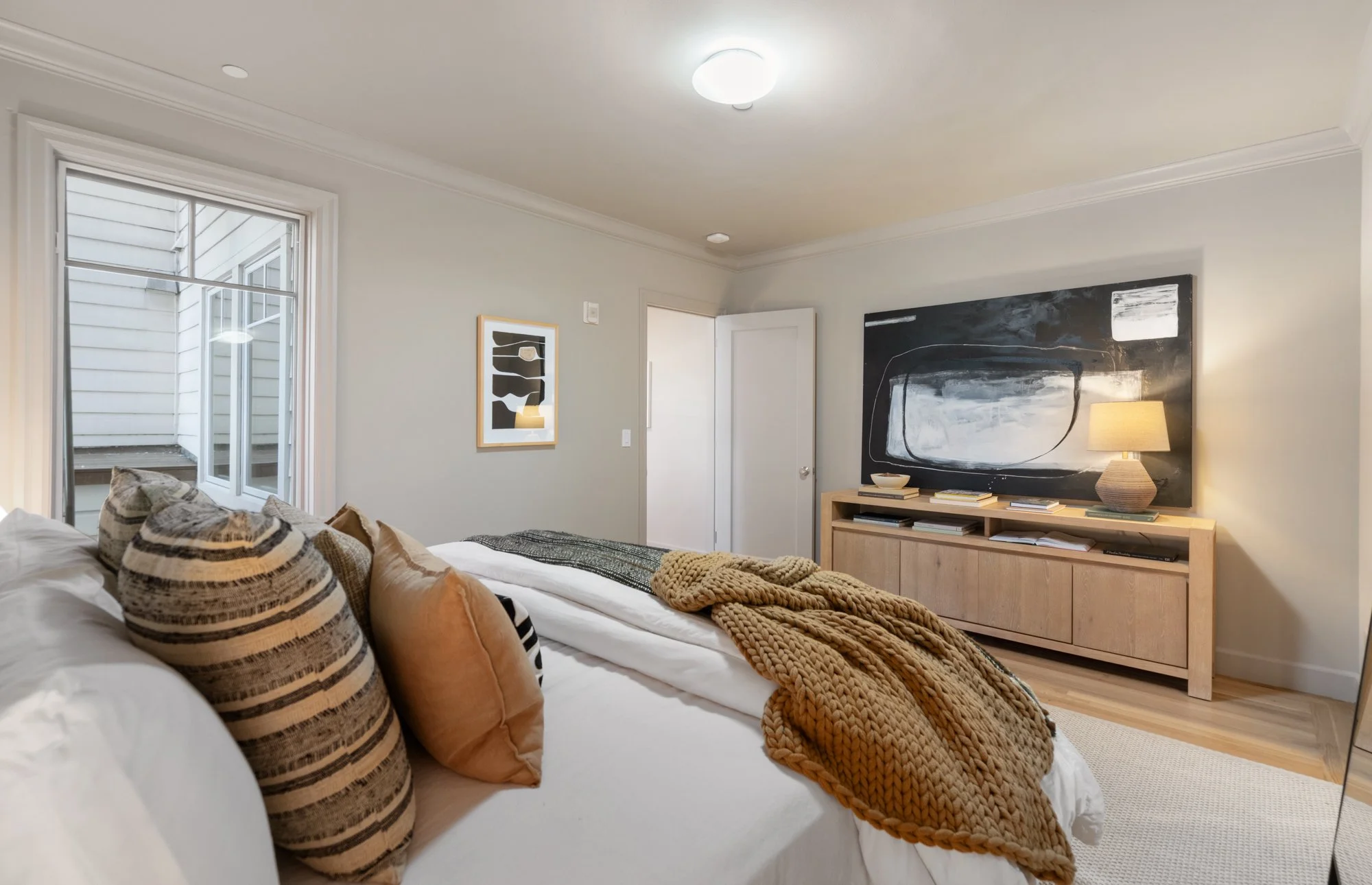 Cozy bedroom with a bed featuring patterned pillows and a chunky knit blanket, artwork on the walls, a wooden media console, a table lamp, and a window with a view of a house exterior.