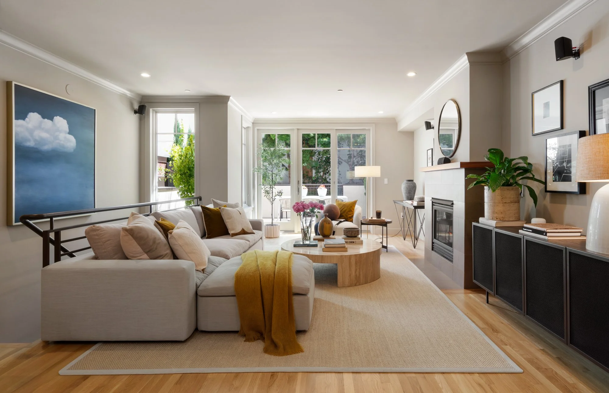 Bright living room with gray couch decorated with beige and yellow pillows, a yellow throw blanket, and a round wooden coffee table with books and vases, white walls with art, and large windows with trees outside.