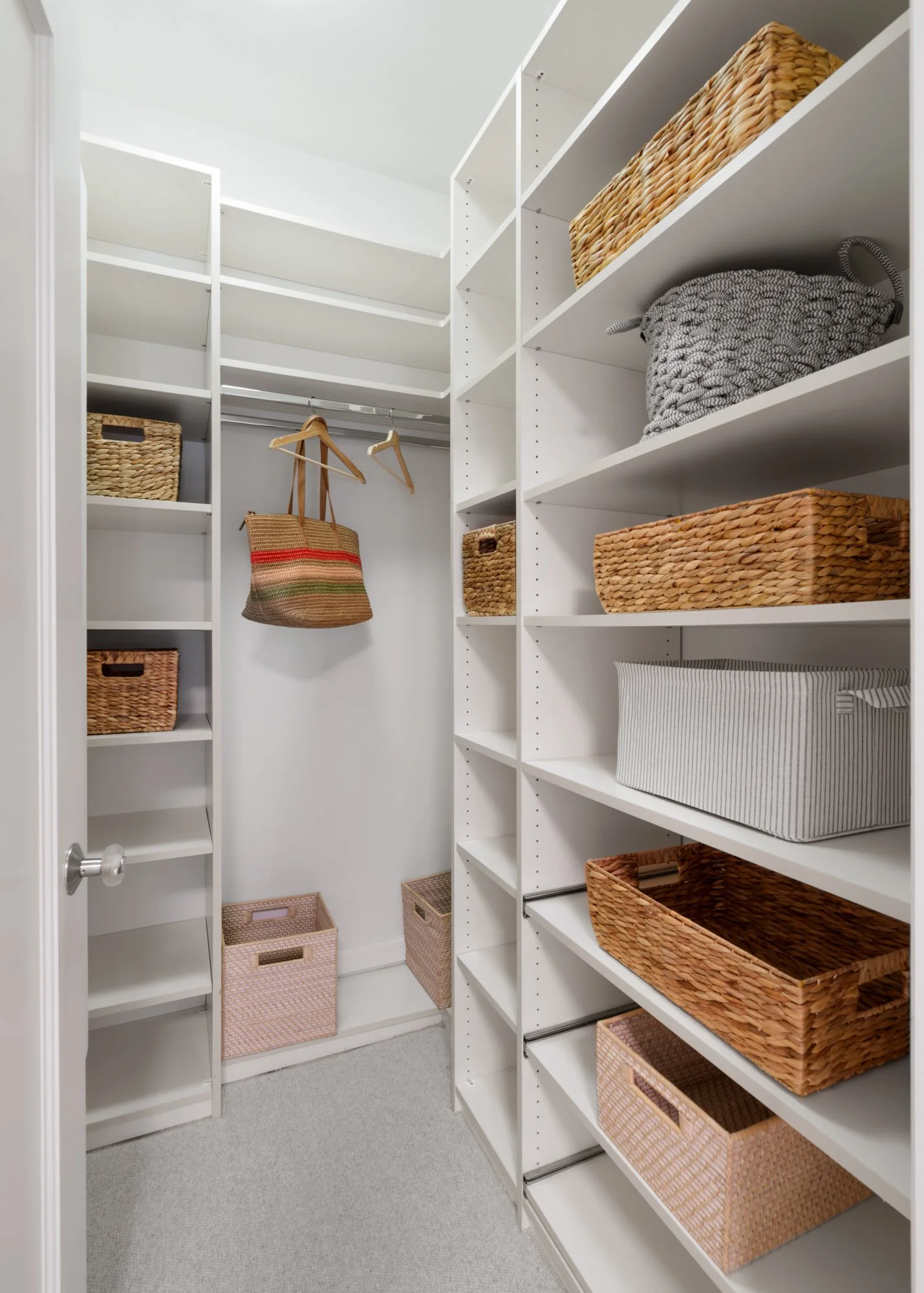 Empty walk-in closet with white shelves and baskets for storage, containing some wicker baskets, cloth bins, and a hanging rod with a tote bag and hangers.