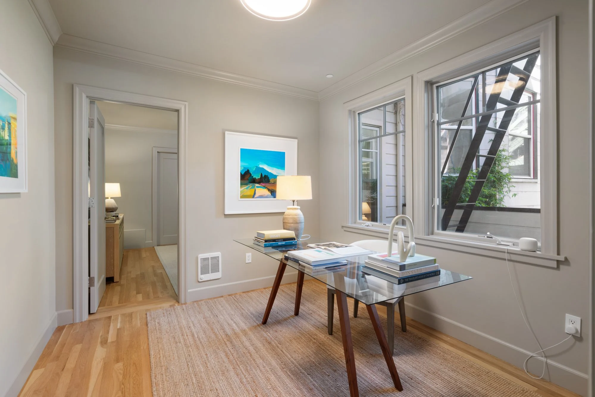 A bright, cozy living space with large windows, light-colored walls, a glass table with books, and a table lamp. Artworks adorn the walls, and a view of outside greenery is visible through the windows.