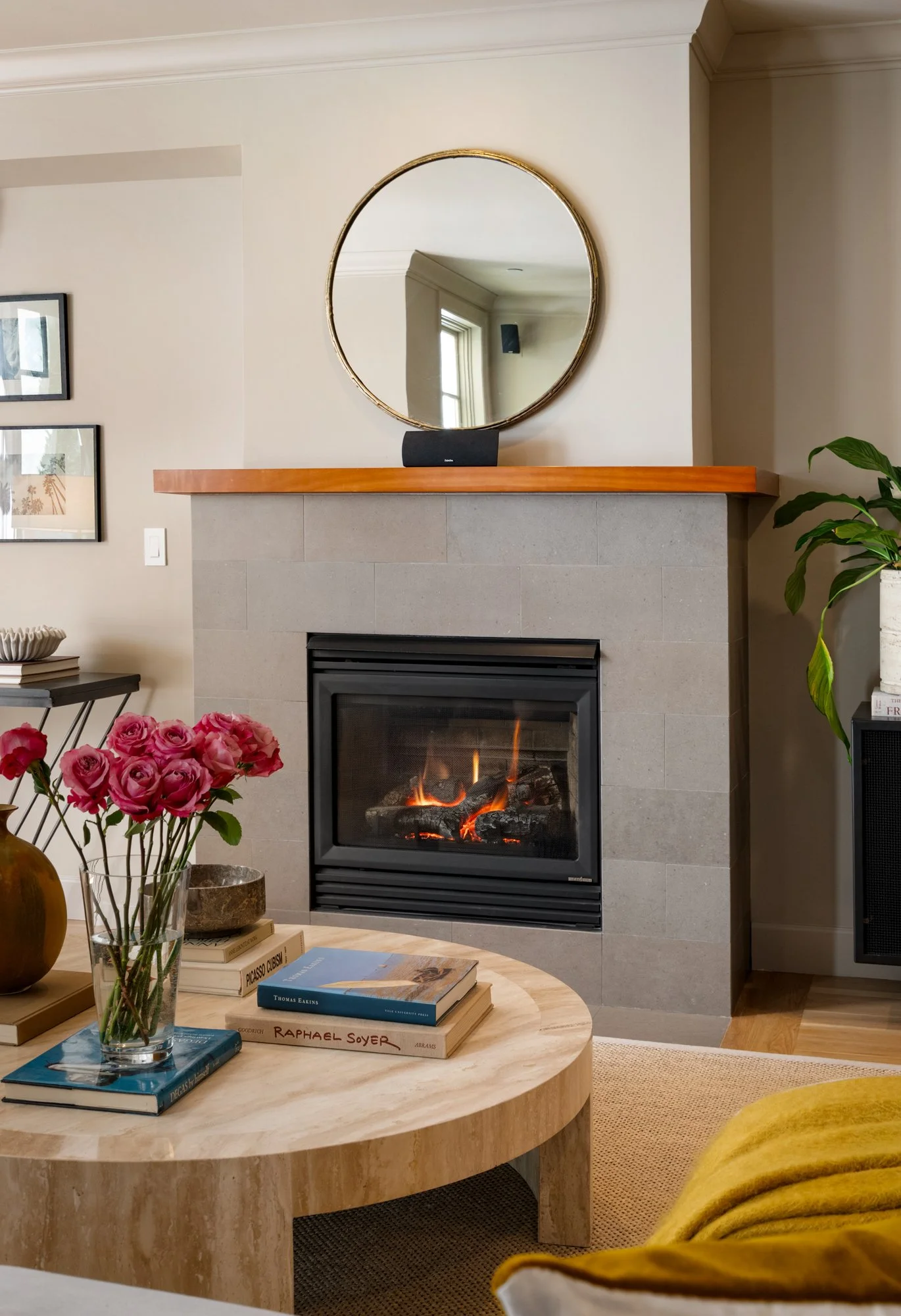 A cozy living room with a fireplace, a round coffee table with books and a vase of pink roses, and a mirror above the fireplace. There is a yellow blanket on a chair or sofa in the foreground and houseplants near the window.