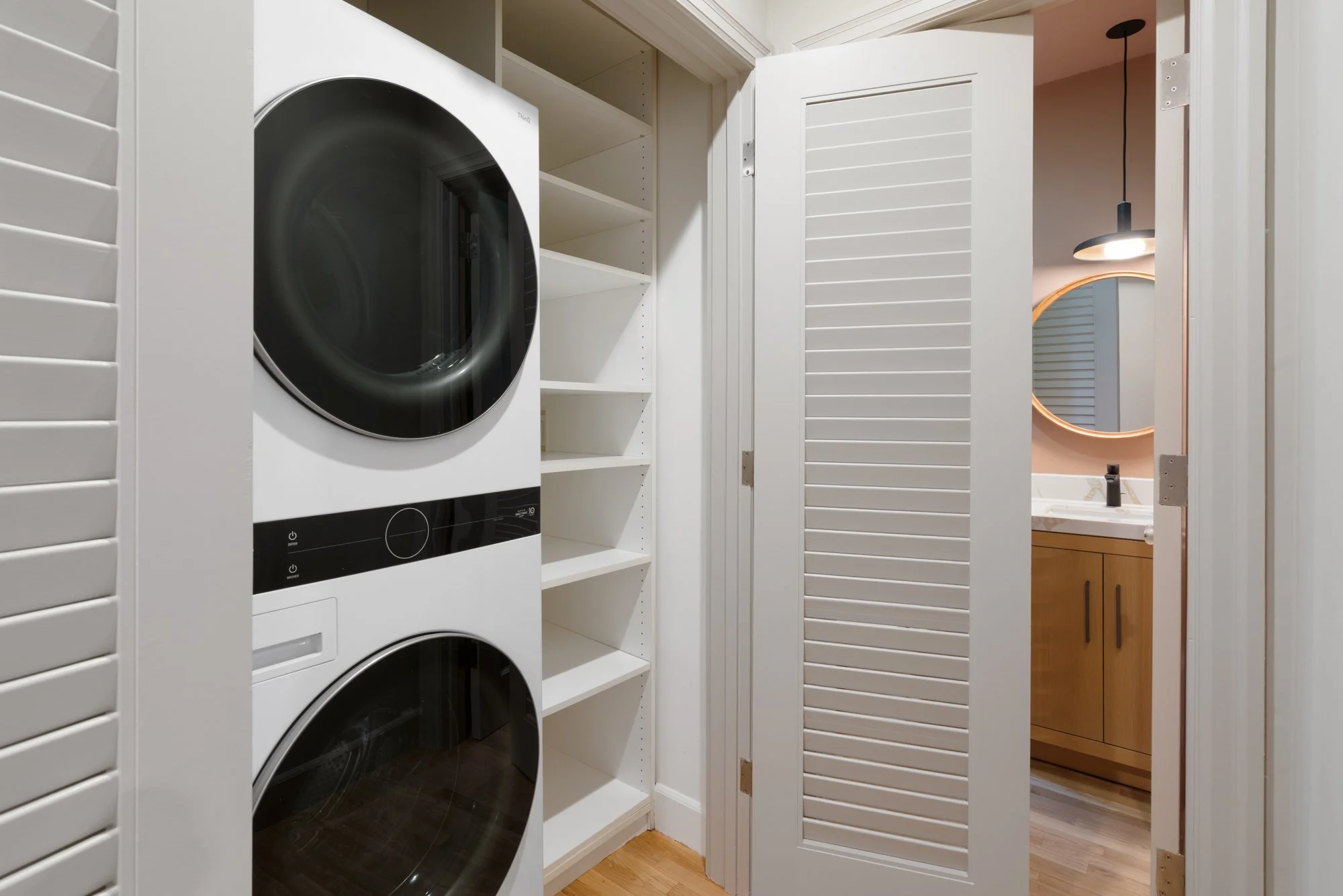 A laundry room with a stacked washer and dryer, white shelves, a louvered closet door, and a bathroom with a mirror and pendant light visible through the open door.