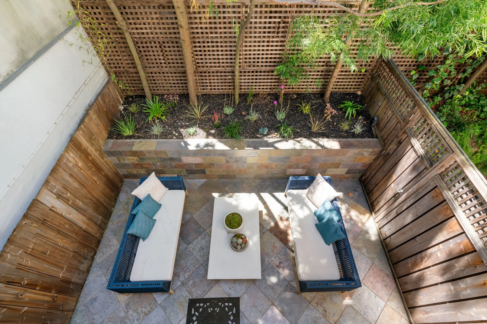 Overhead view of a small outdoor patio with a white table, two wicker chairs with blue cushions, a potted plant, and a wooden privacy fence with surrounding greenery.