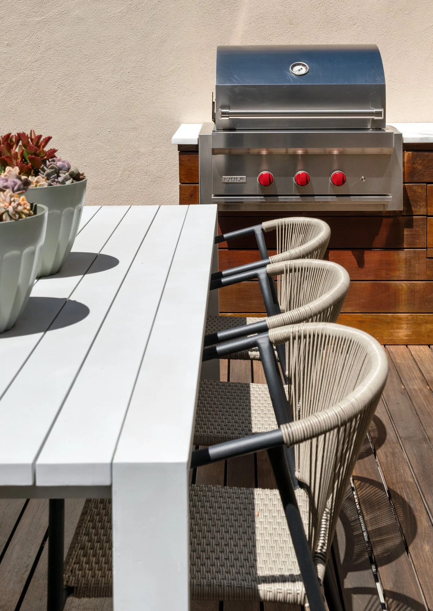 Outdoor patio with white slatted table, beige wicker chairs, potted succulents, and a stainless steel barbecue grill against a beige wall.