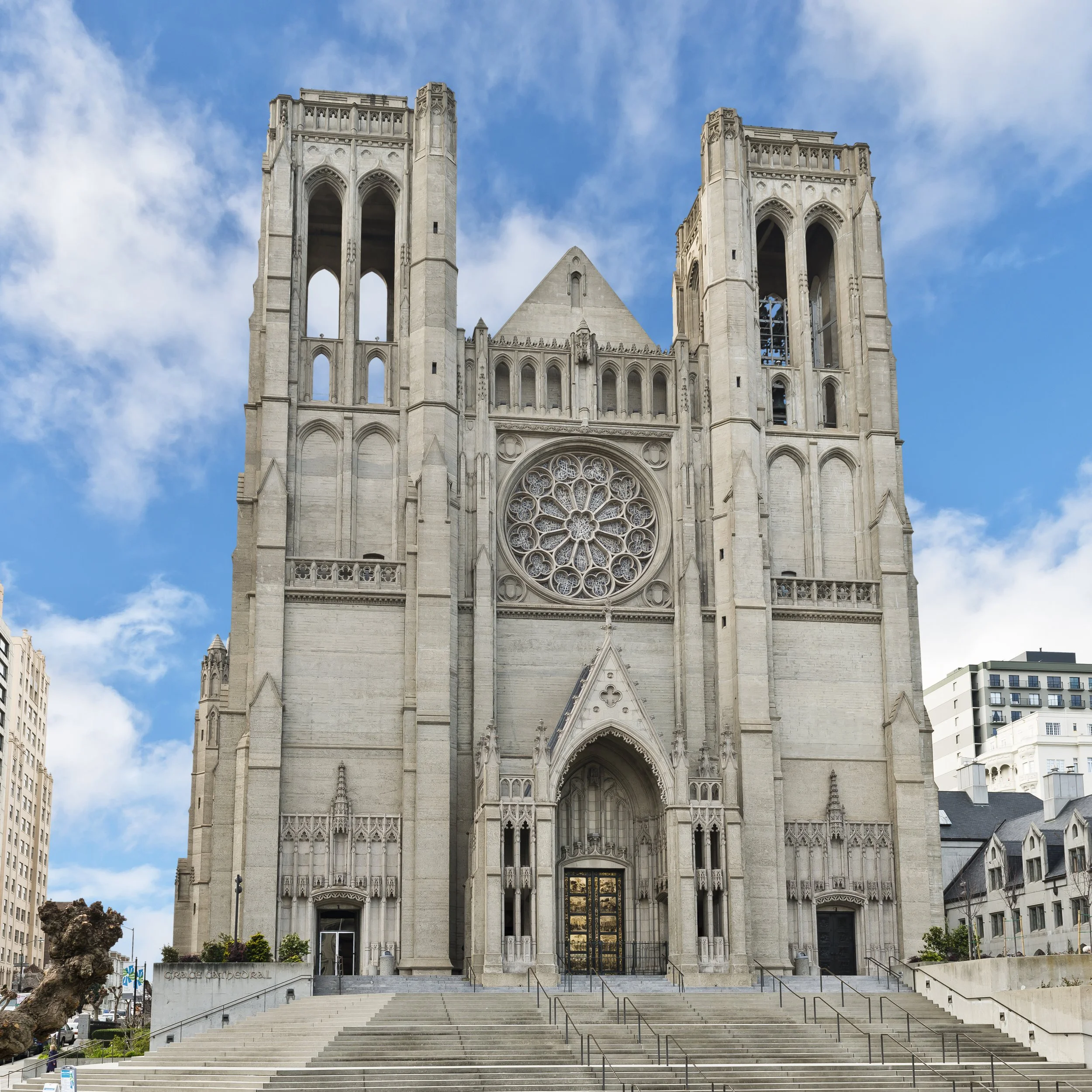 Front view of Grace Cathedral, a Gothic-style church with two tall towers, a large rose window, steps leading up to the entrance, and modern buildings in the background under a partly cloudy sky.
