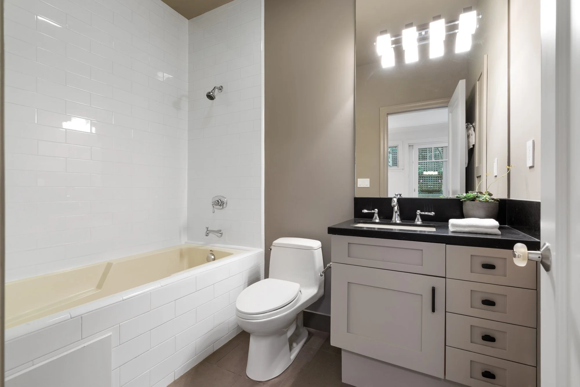 A modern bathroom with white subway tile shower, beige bathtub, white toilet, and a vanity with a black countertop, silver fixtures, and a mirror above it. There is a window with blinds in the background and a potted plant on the counter.