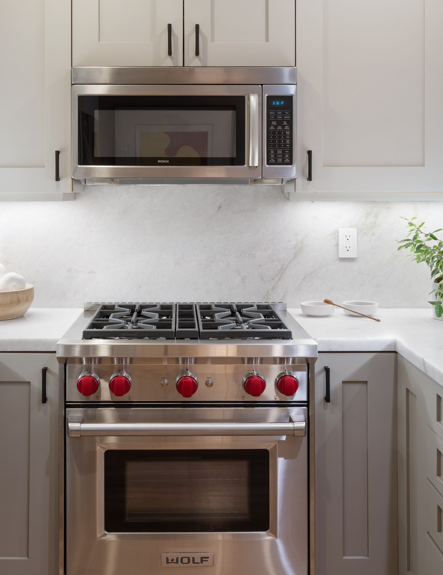 Stainless steel Wolf gas stove with five burners and large oven, white kitchen cabinets with black handles, and a white marble countertop.