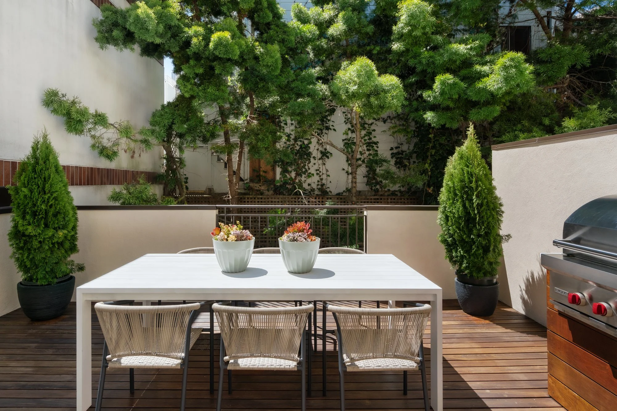 A white outdoor table with five chairs on a wooden deck. Two large potted shrubs flank the table, and there are two white flower pots with succulents on the table. A barbecue grill is on the right. In the background, there's a wooden fence, tall tree