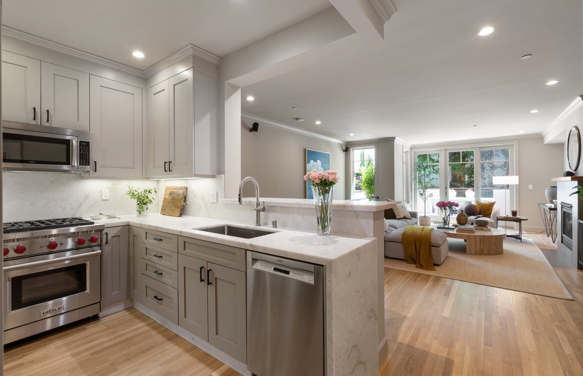A modern kitchen with white cabinets, stainless steel appliances, and a large living room with a sofa, flowers, and large windows letting in natural light.